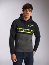 Sweat à capuche Alpinestars Parallel, streetwear décontracté, blocs de couleur noir et gris anthracite, bande jaune vif sur la poitrine avec logo Alpinestars, logo Astars blanc, porté par un mannequin avec un jean bleu, design moderne inspiré du sport automobile