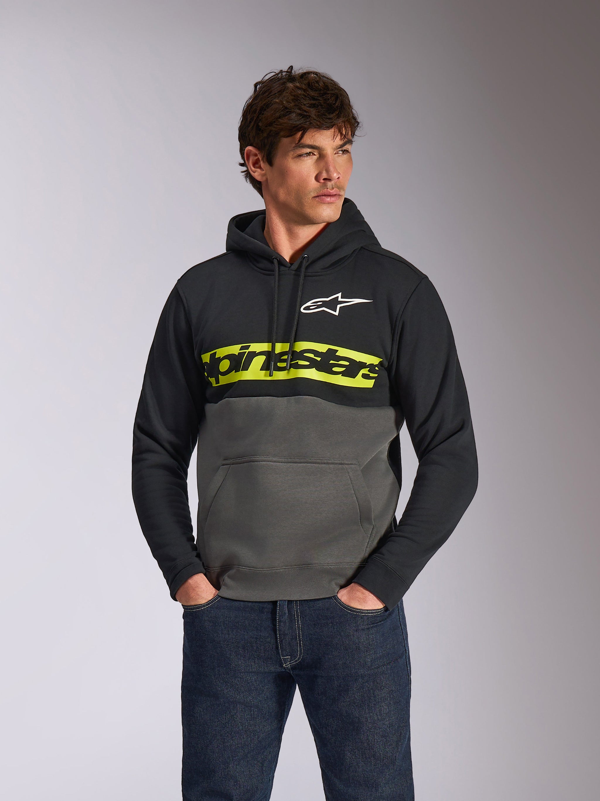 Sweat à capuche Parallel Alpinestars, polaire décontractée avec design en blocs de couleurs, noir et gris anthracite avec une bande jaune vif sur la poitrine, logo Alpinestars textuel et logo étoile blanc, porté par un modèle masculin avec un jean bleu foncé