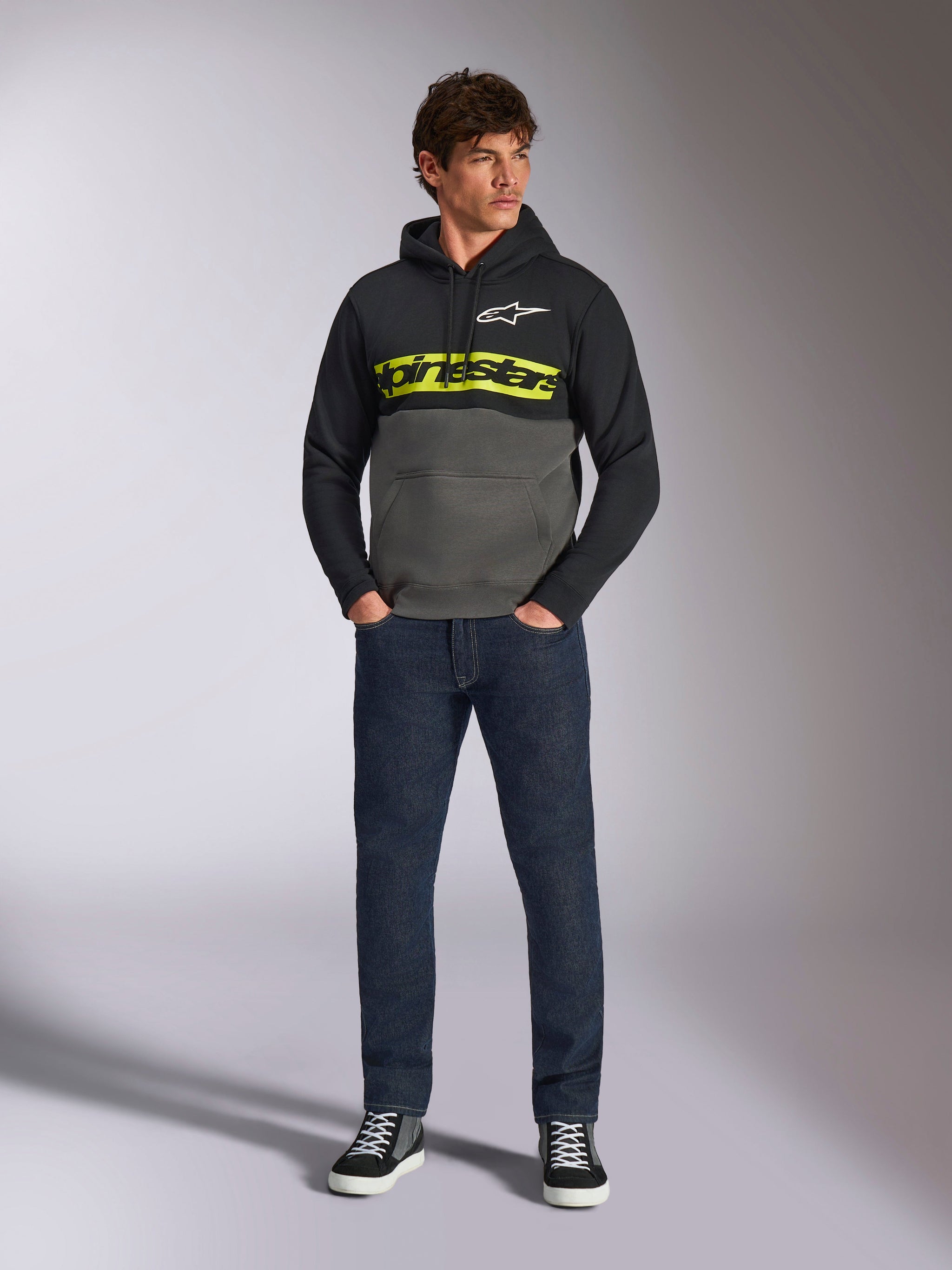 Sweat à capuche Parallel Alpinestars, pull à blocs de couleurs, noir et gris Gunmetal, logo Alpinestars jaune vif et logo étoile blanc sur la poitrine, porté par un mannequin homme avec un jean bleu foncé et des baskets noires, style streetwear motorsport décontracté