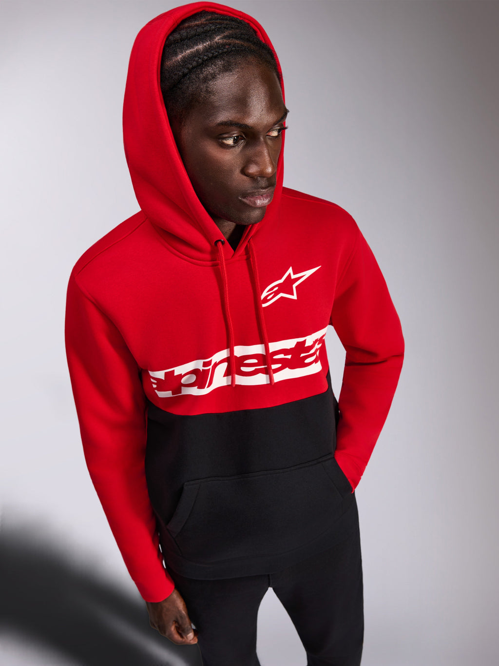 Alpinestars Parallel Hoodie, sweat à capuche décontracté en polaire, design à blocs de couleurs rouge et noir, logo Alpinestars Word et logo Astars blancs sur la poitrine, porté par un homme avec la capuche relevée et un pantalon noir, streetwear d'inspiration sport mécanique