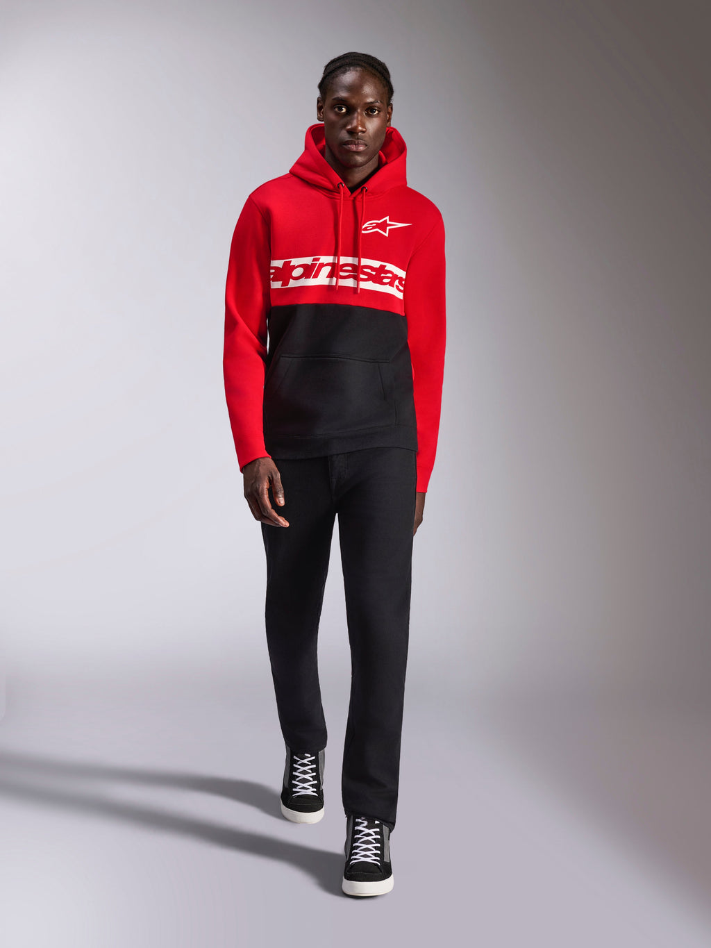 Sweat à capuche Parallel Alpinestars, polaire à enfiler en blocs de couleurs rouge et noir, logo textuel Alpinestars blanc sur la poitrine et logo Astars sur la poitrine gauche, porté par un mannequin avec un pantalon noir et des baskets montantes, style streetwear décontracté