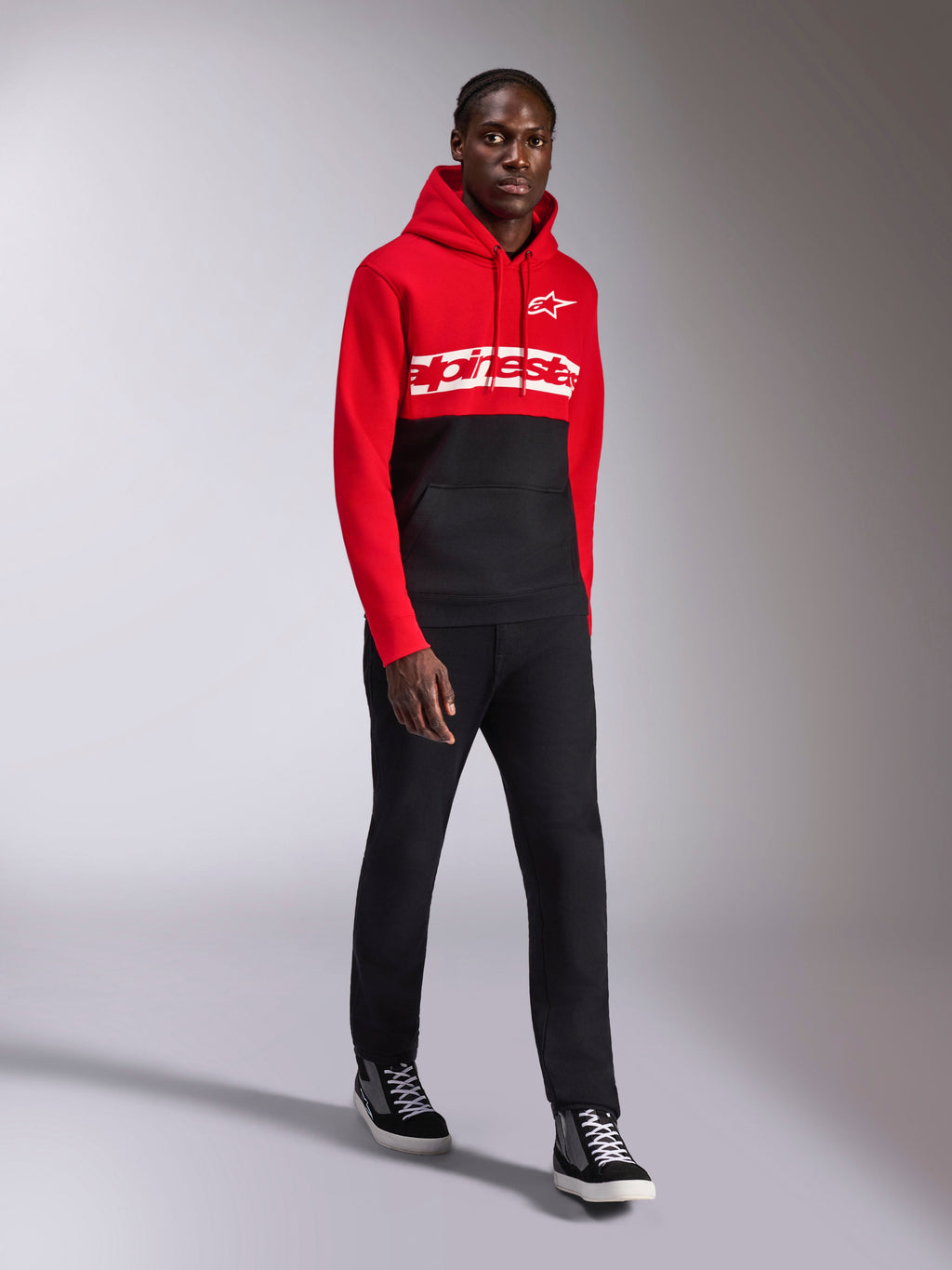 Sweat à capuche Alpinestars Parallel, sportswear décontracté, design à blocs de couleurs rouge et noir, avec lettrage blanc et logo étoilé Alpinestars sur la poitrine, porté avec un pantalon noir et des baskets, look moderne inspiré des sports mécaniques