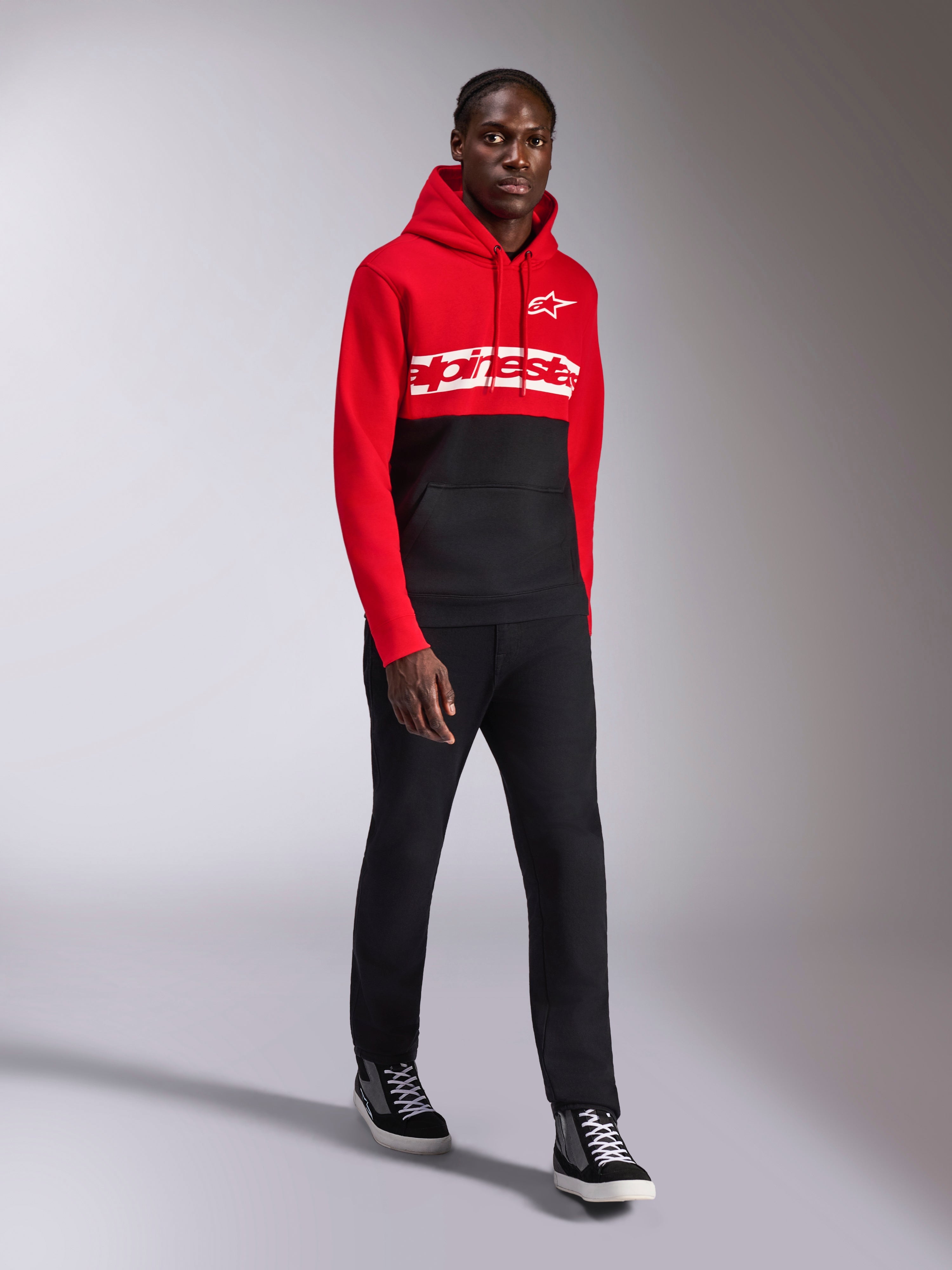 Sweat à capuche Alpinestars Parallel, sportswear décontracté, design à blocs de couleurs rouge et noir, avec lettrage blanc et logo étoilé Alpinestars sur la poitrine, porté avec un pantalon noir et des baskets, look moderne inspiré des sports mécaniques