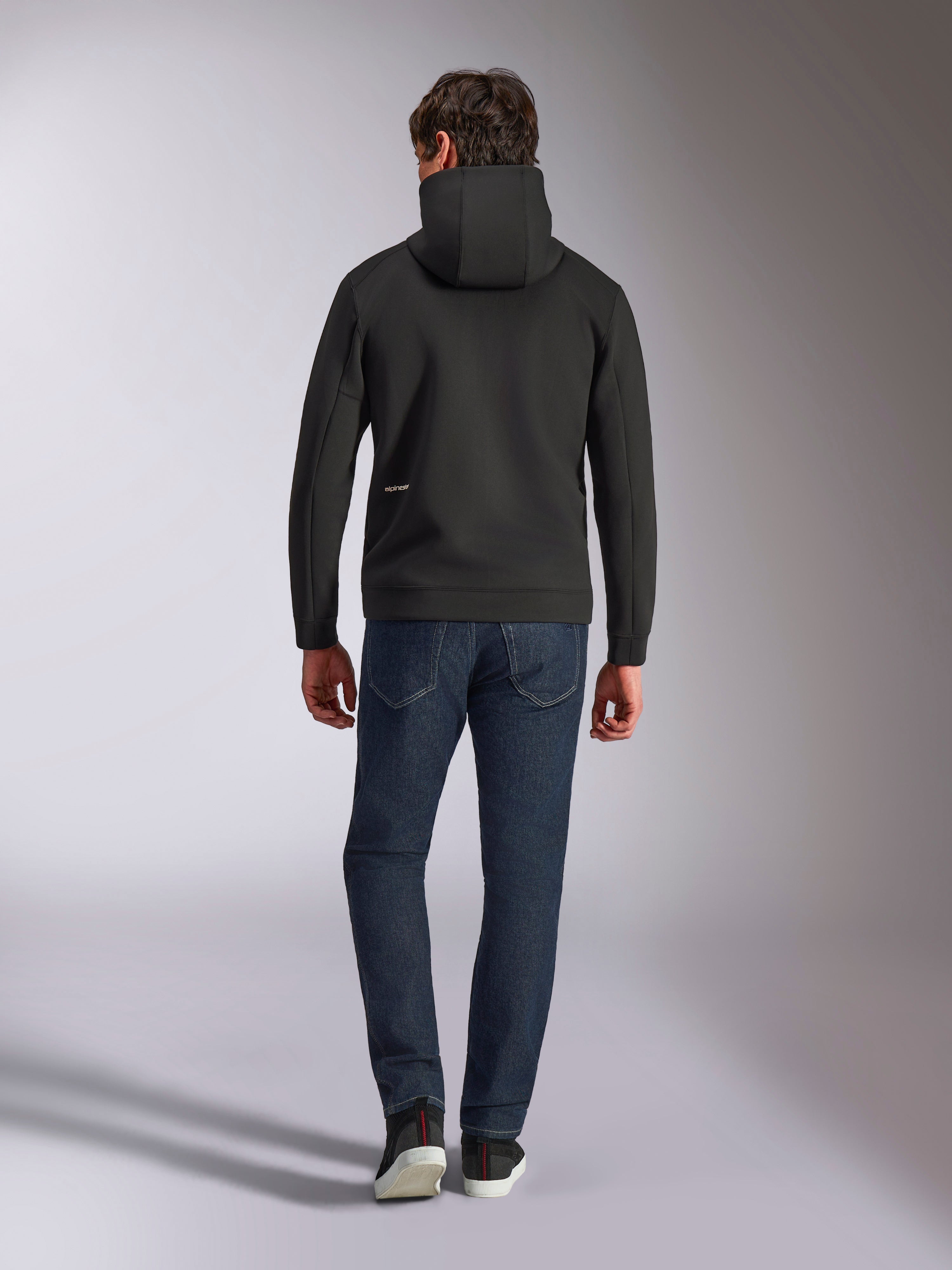 Alpinestars Cypher Hoodie, black double-face fleece, back view showing a small white Alpinestars logo on the lower left hip, high-neck hood, paired with dark blue jeans, casual motorsport-inspired streetwear