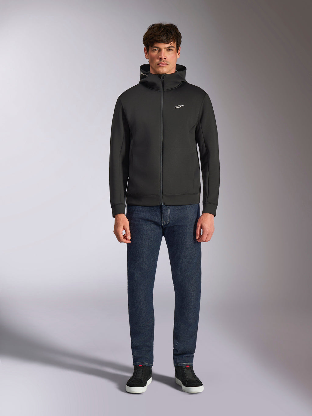 Model wearing the Alpinestars Cypher Hoodie, Black zip-front fleece hoodie, high-neck hood with a small white Astars logo on the chest, paired with dark denim jeans and black sneakers, casual motorsport-inspired streetwear