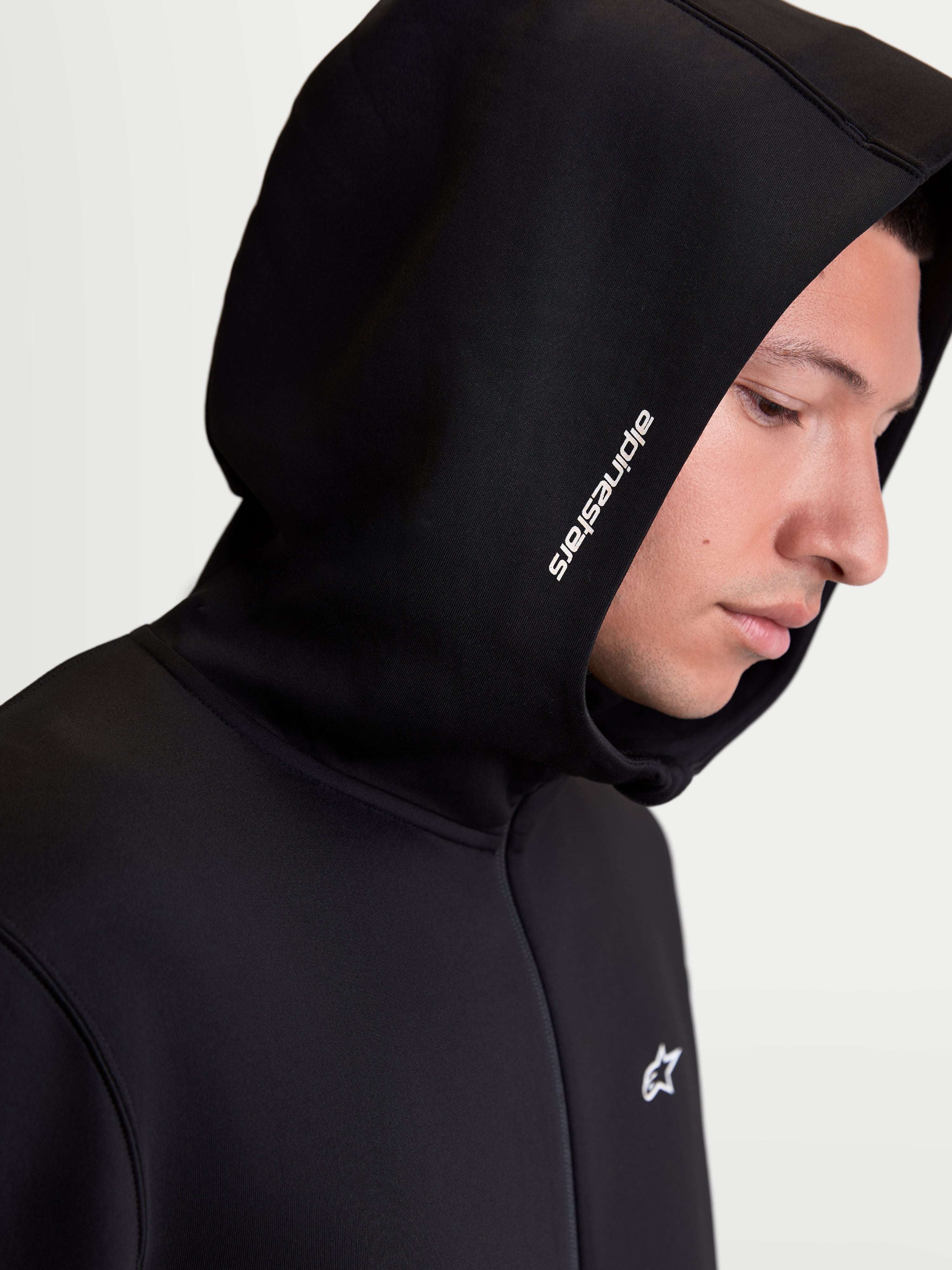 Alpinestars sweat à capuche Cypher, sweat à capuche sportswear zippé en polaire pour homme, noir, vue rapprochée sur modèle montrant la capuche à col haut avec logo Alpinestars blanc et polaire technique tri-mélange