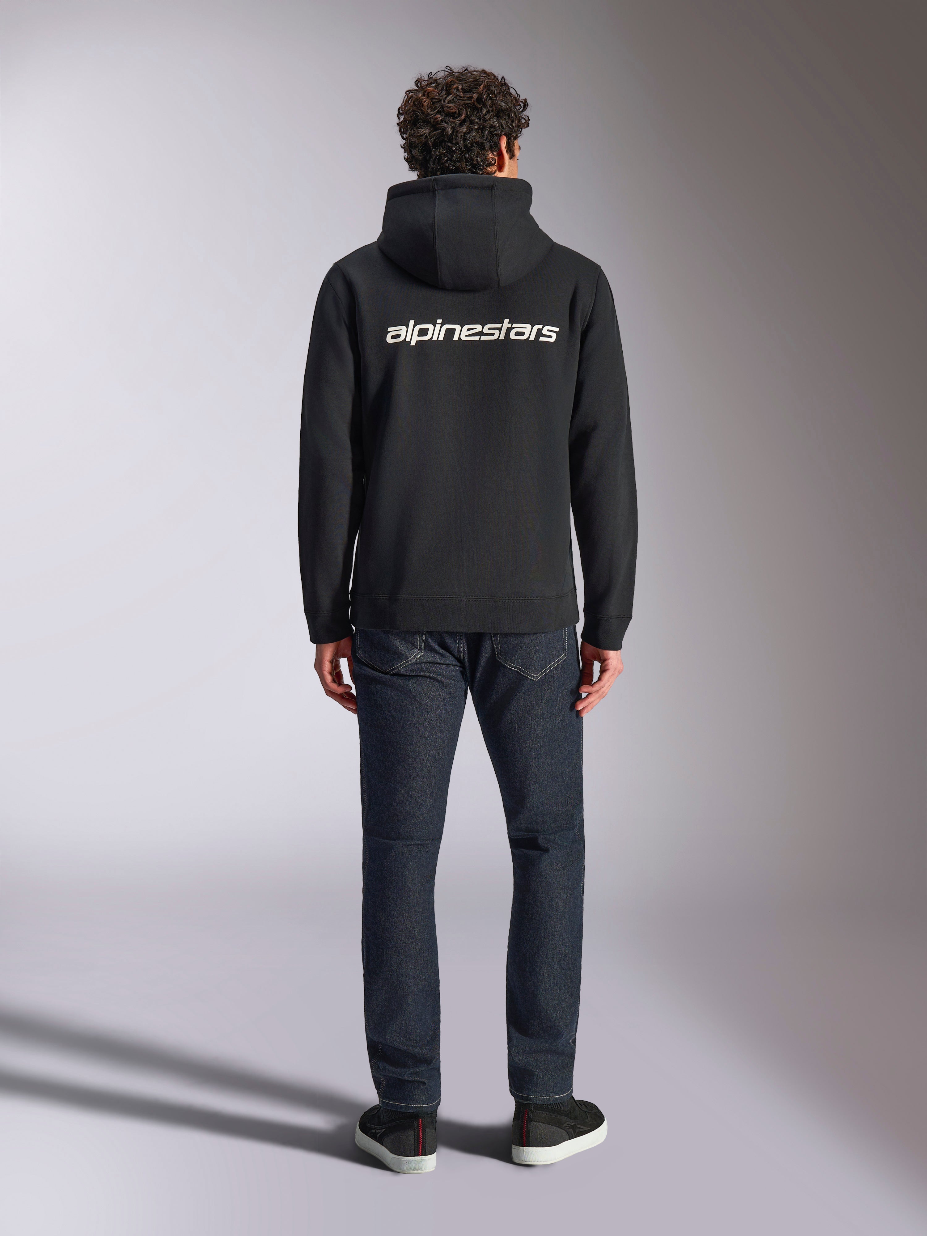 Alpinestars Transverse Hoodie, casual motorsport streetwear, Black with white Alpinestars word logo on the back, long sleeves, ribbed hem and cuffs, worn from behind with dark denim jeans