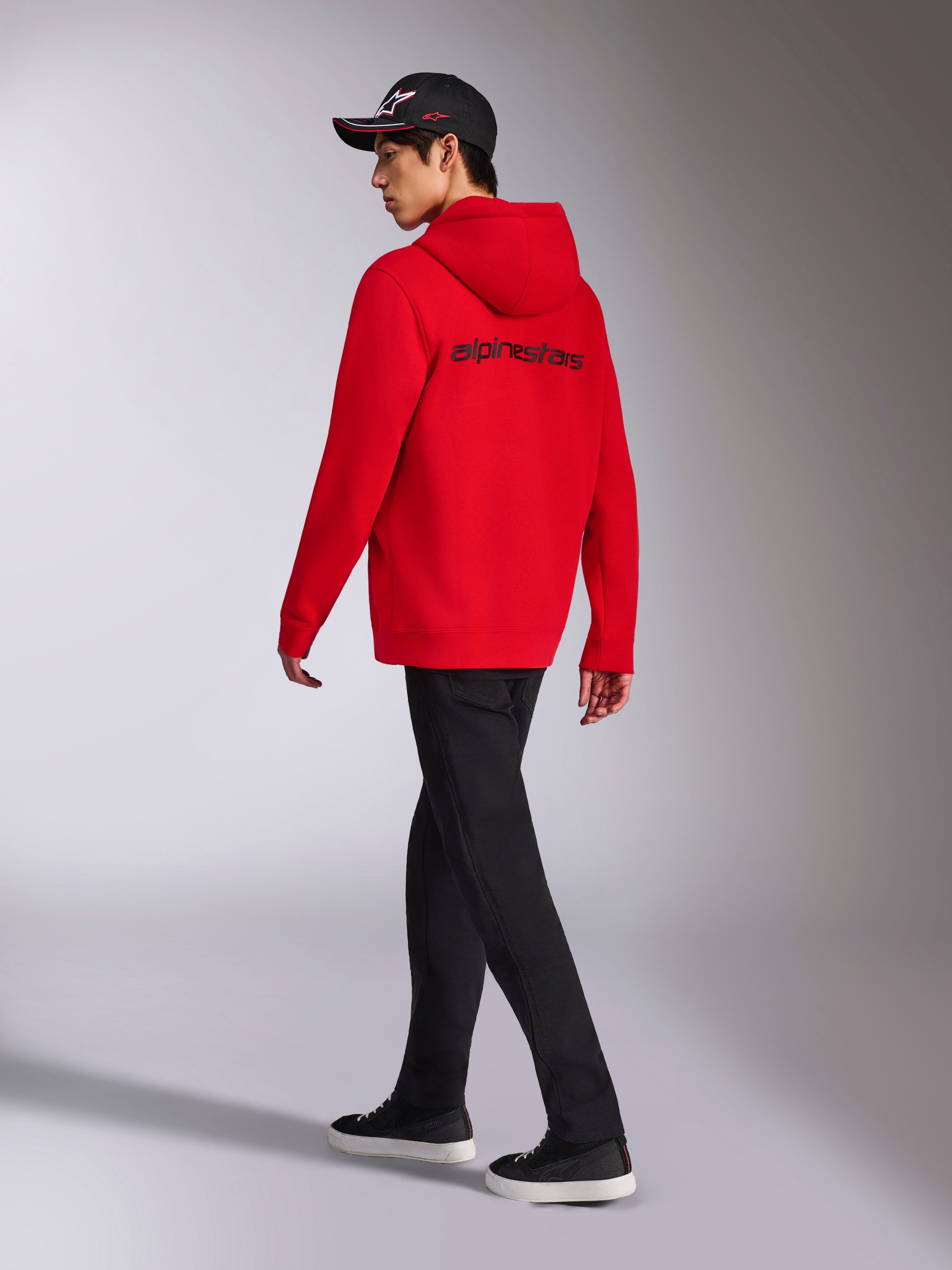 Sweat à capuche Transverse Alpinestars, sweat à capuche en polaire rouge zippé, vue de dos présentant le logo Alpinestars noir, porté par un mannequin avec un pantalon noir et une casquette à logo noire, streetwear décontracté inspiré par les sports mécaniques