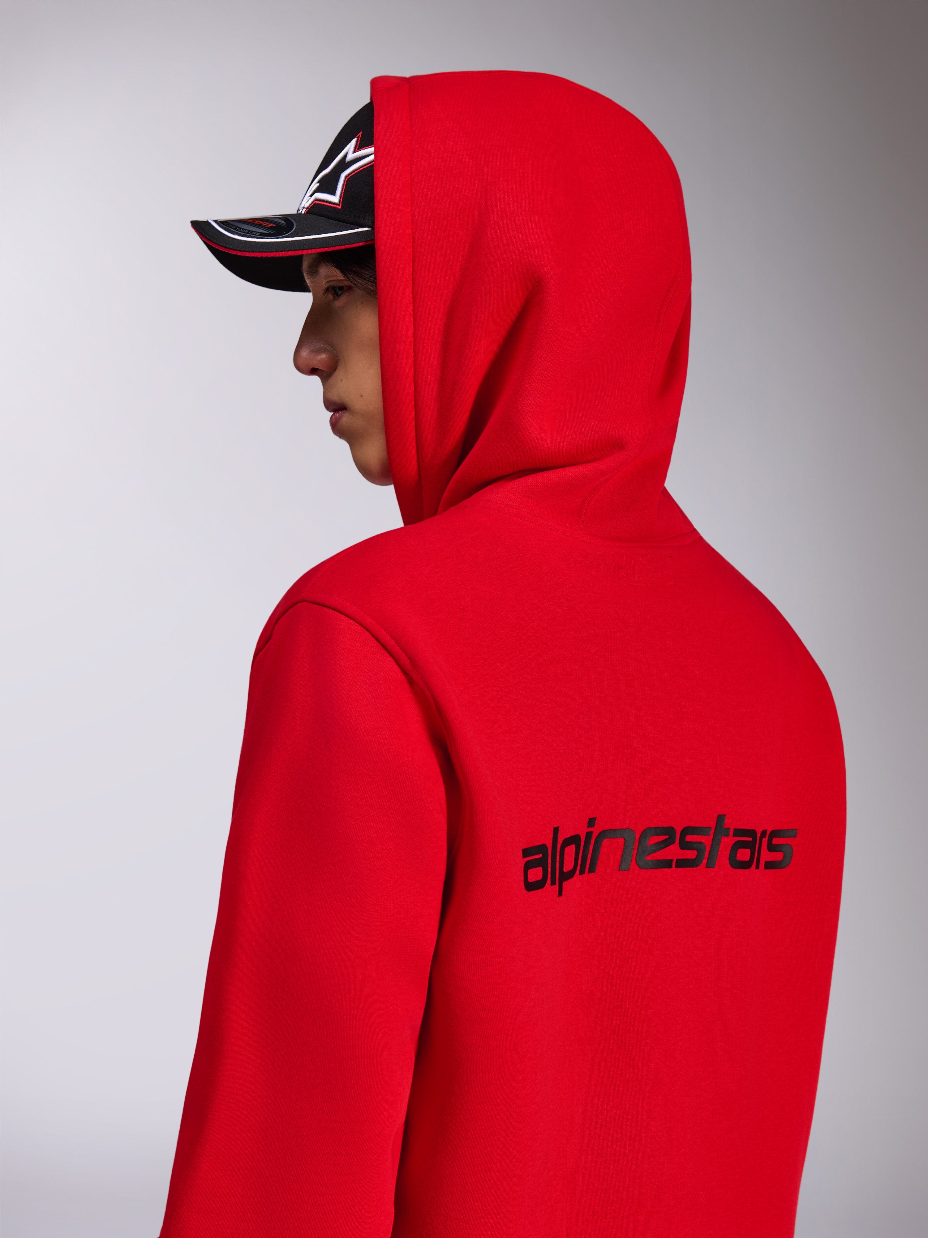 Sweat à capuche Alpinestars Transverse, polaire rouge à capuche, vue de profil arrière montrant le logo Alpinestars noir dans le dos, porté avec la capuche relevée et une casquette noire, style streetwear décontracté inspiré du sport automobile