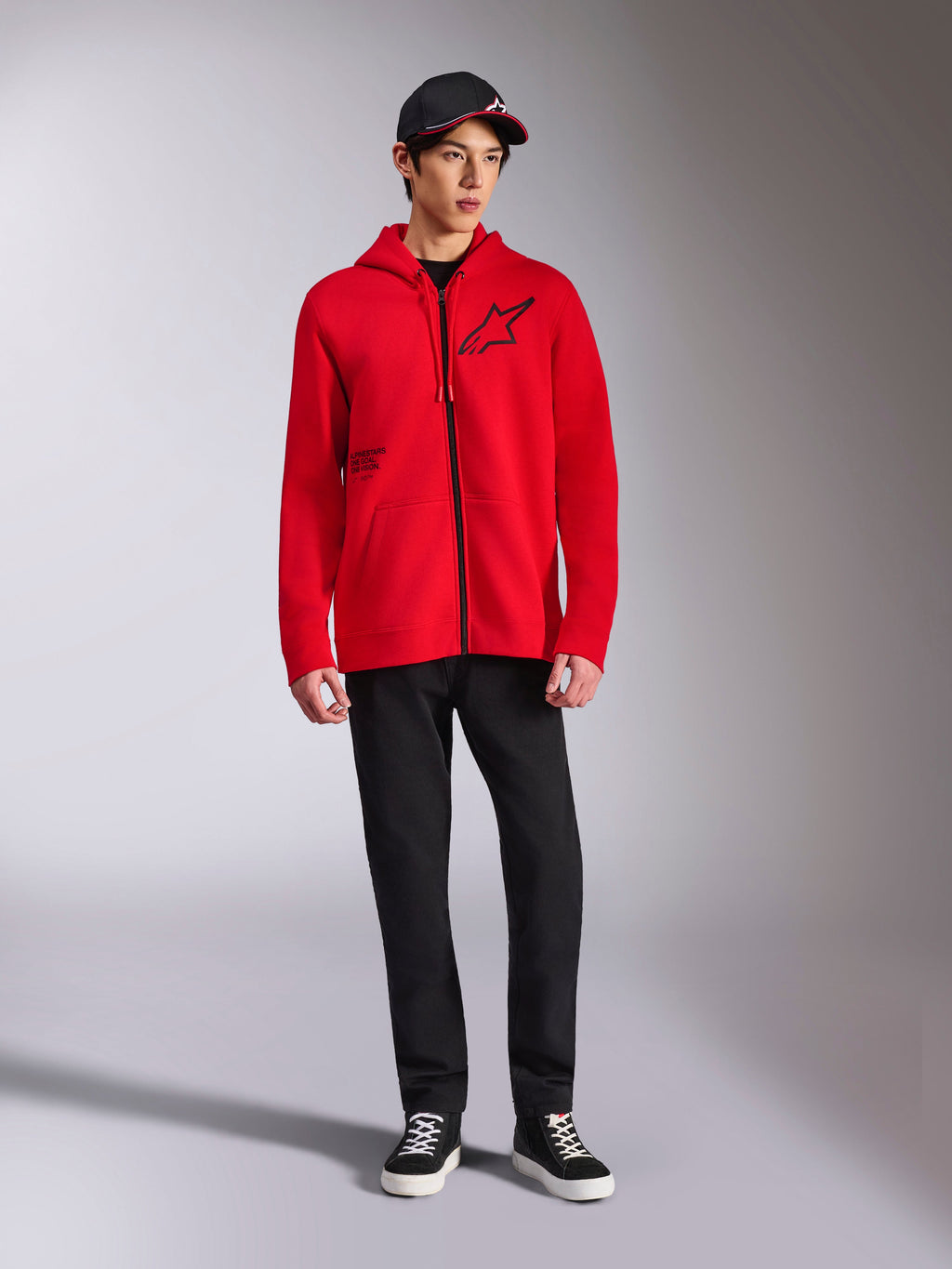 Alpinestars Transverse Hoodie, sweat à capuche rouge en polaire avec fermeture éclair intégrale, logo Astars noir sur la poitrine et imprimé One Goal sur le côté, porté par un mannequin homme avec pantalon noir et casquette, style streetwear décontracté inspiré du sport automobile