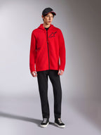 Alpinestars Transverse Hoodie, sweat à capuche rouge en polaire avec fermeture éclair intégrale, logo Astars noir sur la poitrine et imprimé One Goal sur le côté, porté par un mannequin homme avec pantalon noir et casquette, style streetwear décontracté inspiré du sport automobile