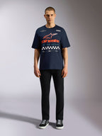 T-shirt Alpinestars Aplomb Oversized CSF, t-shirt à manches courtes bleu marine, grand imprimé sur la poitrine avec logo en dégradé orange et rouge et motif à damier, porté par un mannequin avec un pantalon noir et des baskets, style streetwear sport mécanique décontracté