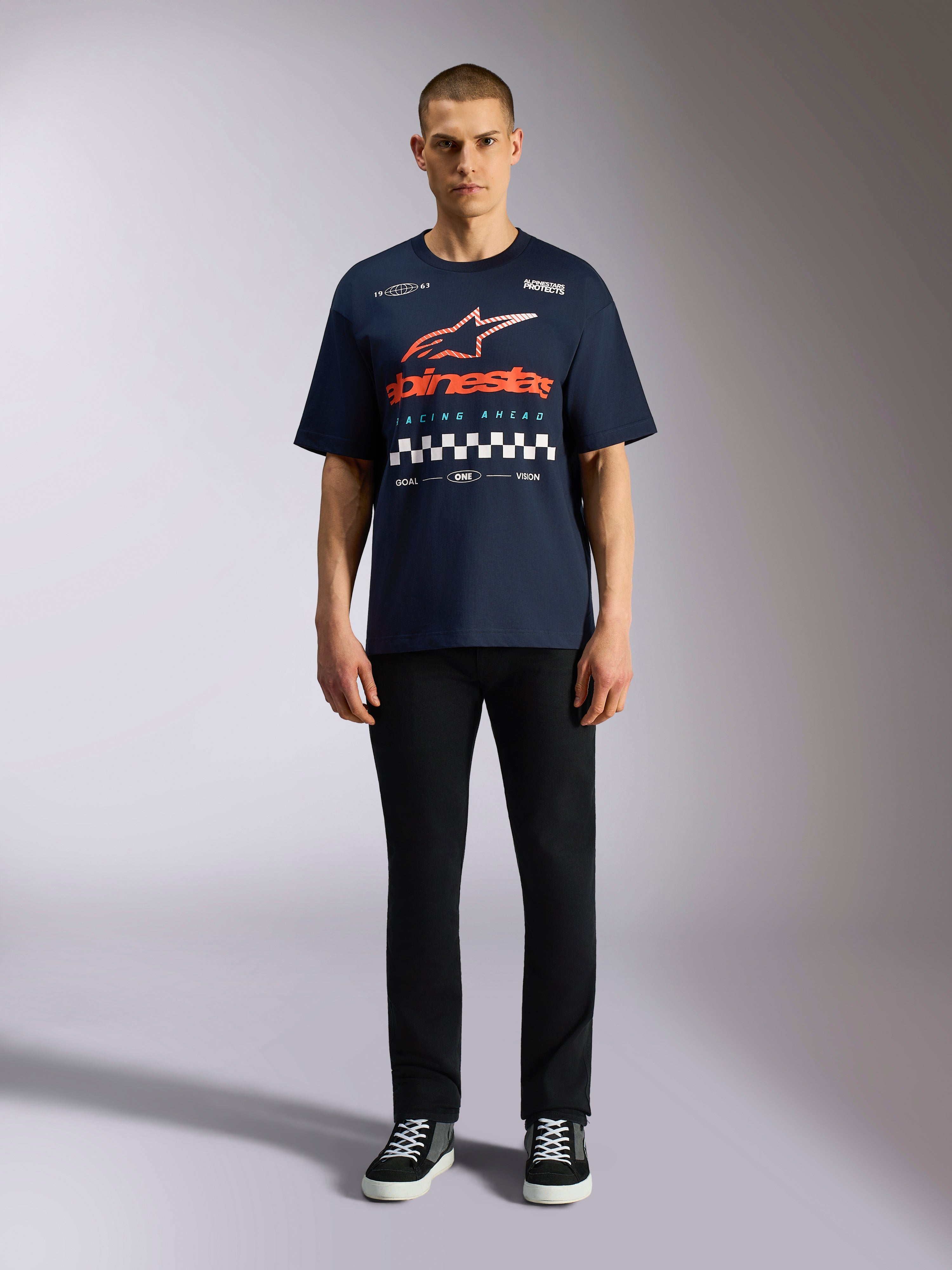 T-shirt Alpinestars Aplomb Oversized CSF, t-shirt à manches courtes bleu marine, grand imprimé sur la poitrine avec logo en dégradé orange et rouge et motif à damier, porté par un mannequin avec un pantalon noir et des baskets, style streetwear sport mécanique décontracté