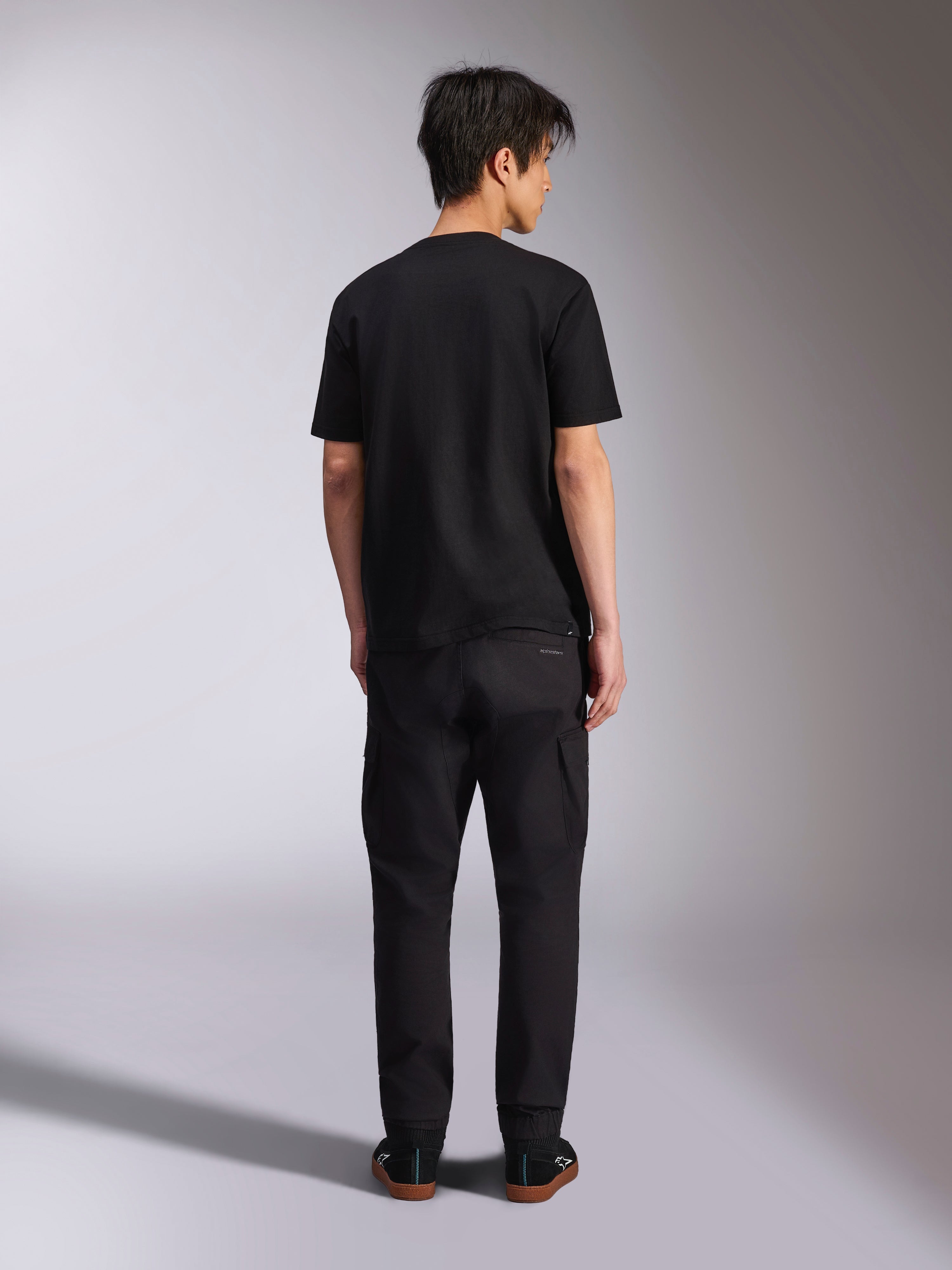 T-shirt Alpinestars Noka CSF, t-shirt à manches courtes, noir, vue de dos du mannequin portant le t-shirt avec un pantalon noir et des baskets, style streetwear décontracté motorsport