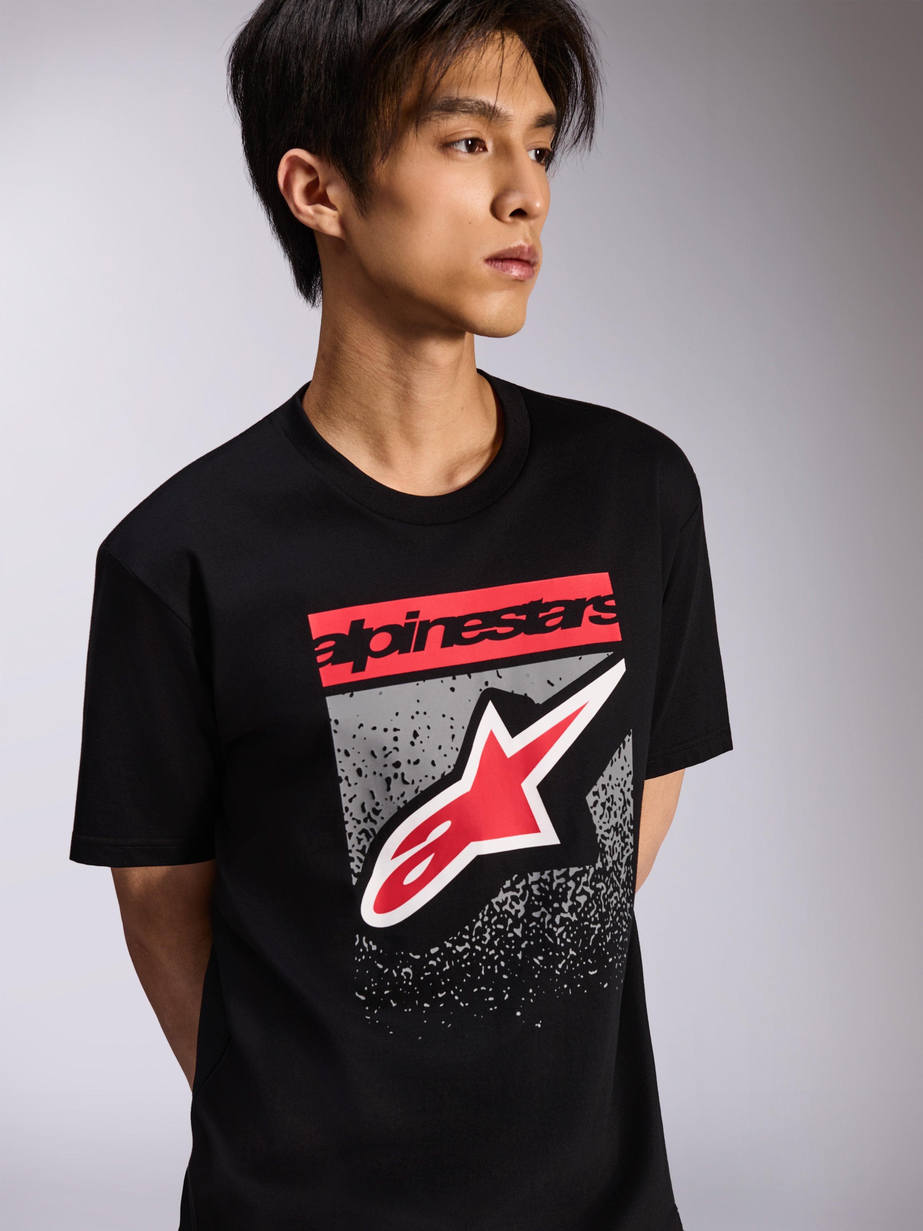 T-shirt Alpinestars Noka CSF, t-shirt à manches courtes, noir, arborant un bloc de logo rouge et un grand logo Astars rouge et blanc sur un fond texturé gris, style streetwear décontracté