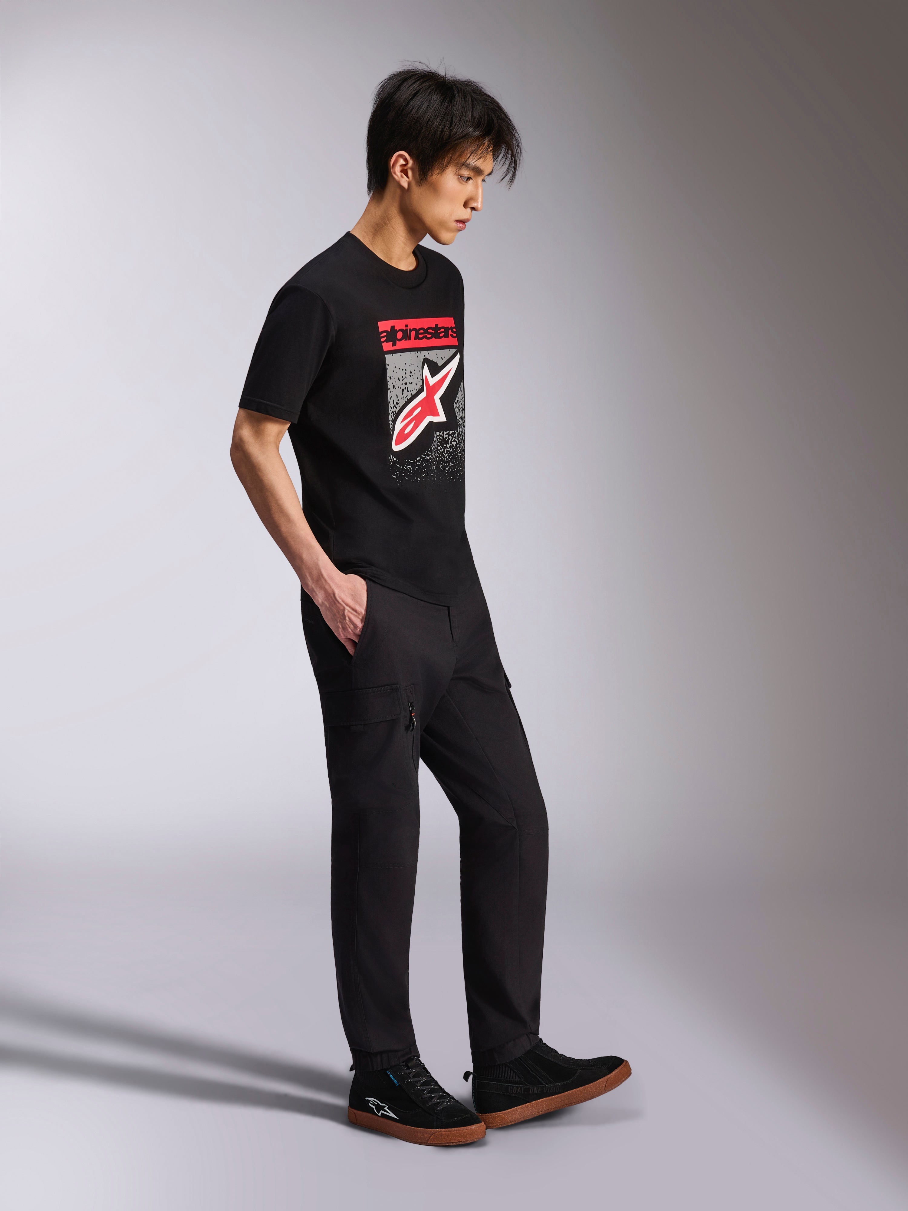 T-shirt Alpinestars Noka CSF, t-shirt à manches courtes, noir, logo Astars à l'avant avec dégradé MX rouge et blanc, porté avec un pantalon cargo noir et des baskets, streetwear décontracté inspiré des sports mécaniques