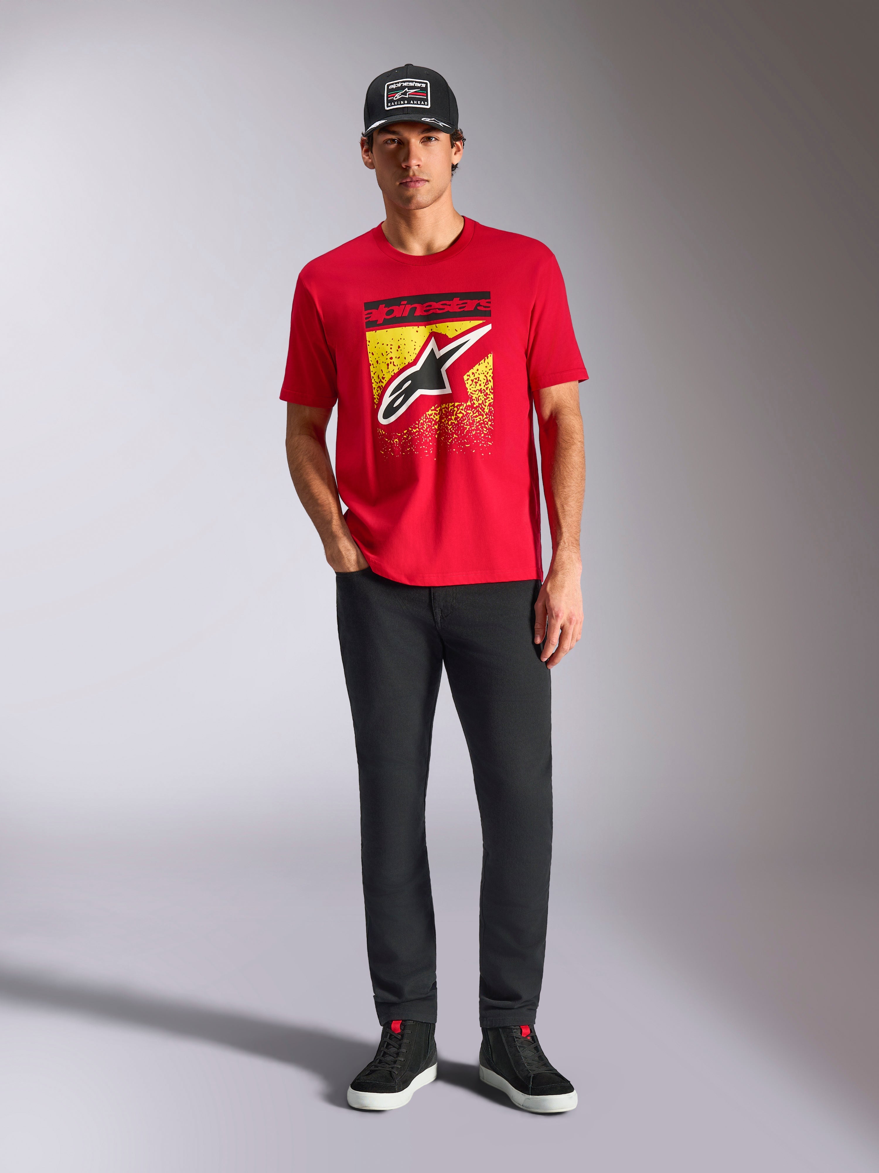T-shirt Alpinestars Noka CSF, t-shirt à manches courtes, rouge, avec un grand logo Astars à l'avant et un dégradé MX jaune et noir, porté avec un pantalon noir et une casquette noire, style streetwear motorsport décontracté
