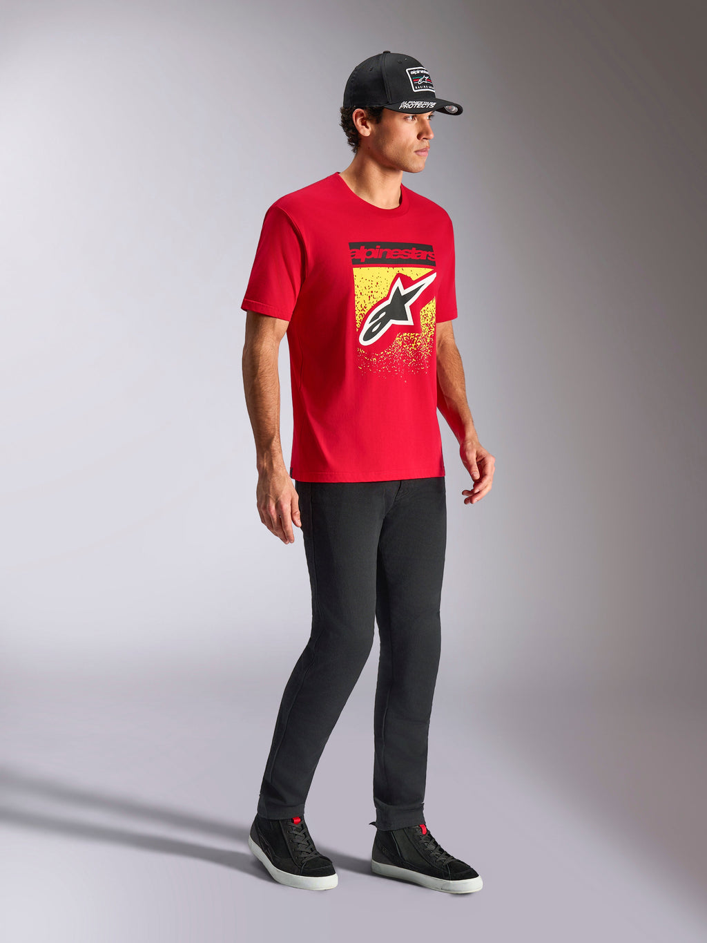 T-shirt Alpinestars Noka CSF, t-shirt à manches courtes, rouge, avec le logo Astars à l'avant et un dégradé MX sur un graphisme jaune, porté avec une casquette Alpinestars noire et un pantalon noir, style streetwear motorsport décontracté