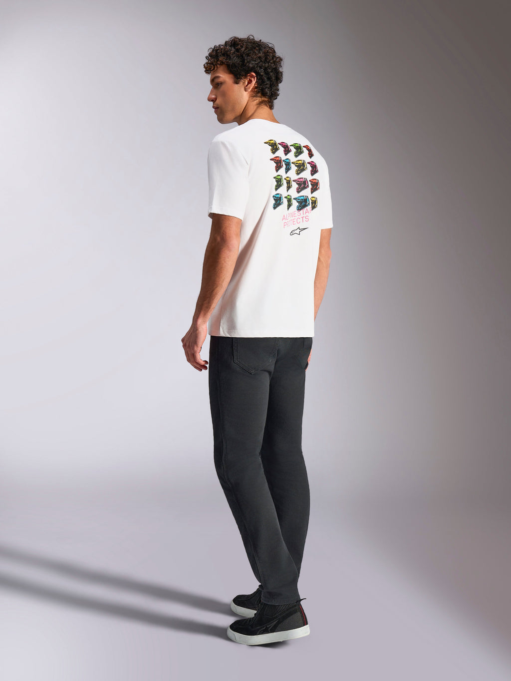 T-shirt Alpinestars Pop Helmet CSF, t-shirt à manches courtes, blanc, vue de dos sur un mannequin présentant un graphisme coloré du casque M10, porté avec un pantalon sombre et des baskets noires, streetwear décontracté inspiré des sports mécaniques