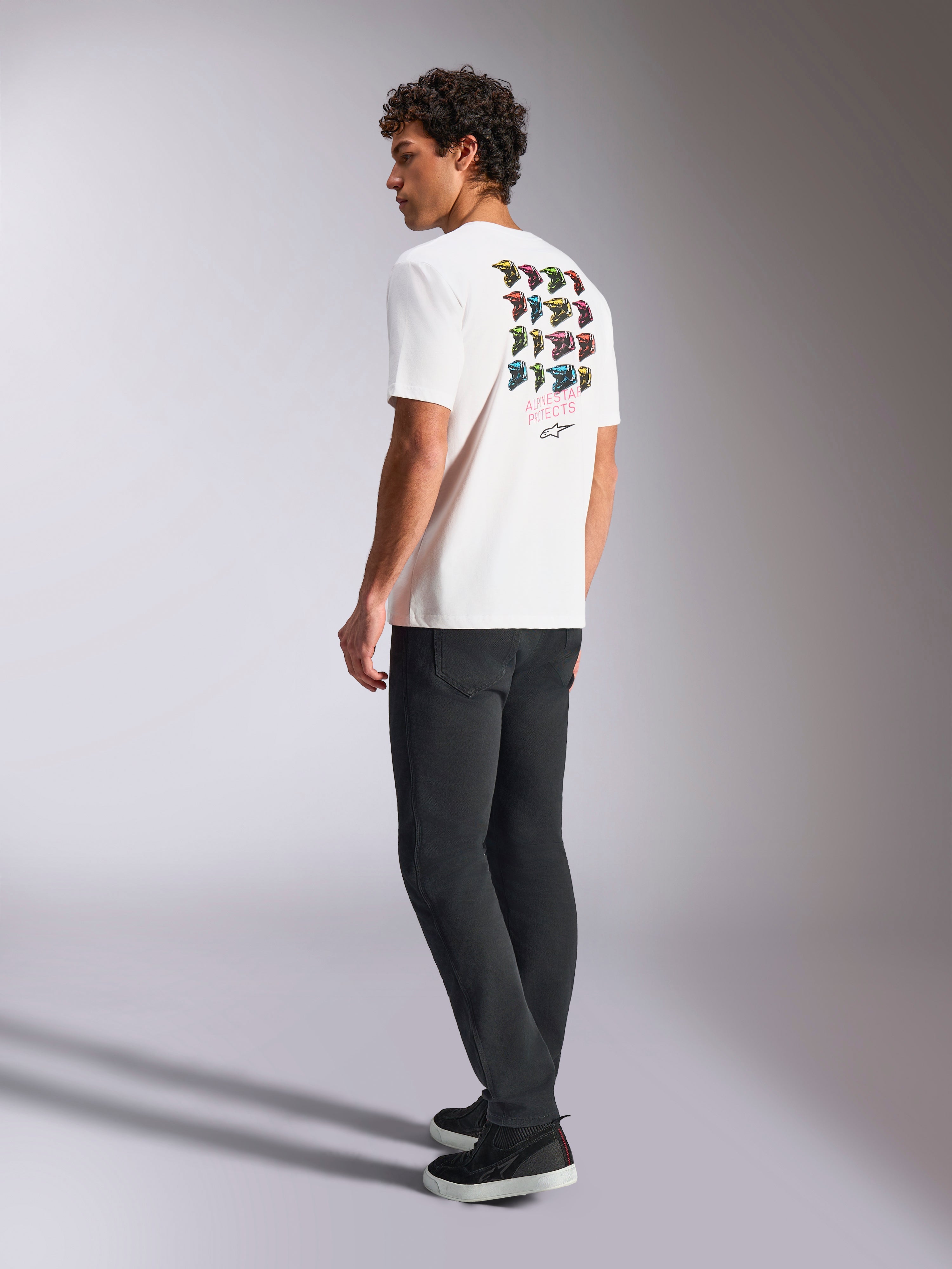 T-shirt Alpinestars Pop Helmet CSF, t-shirt à manches courtes, blanc, vue de dos sur un mannequin présentant un graphisme coloré du casque M10, porté avec un pantalon sombre et des baskets noires, streetwear décontracté inspiré des sports mécaniques