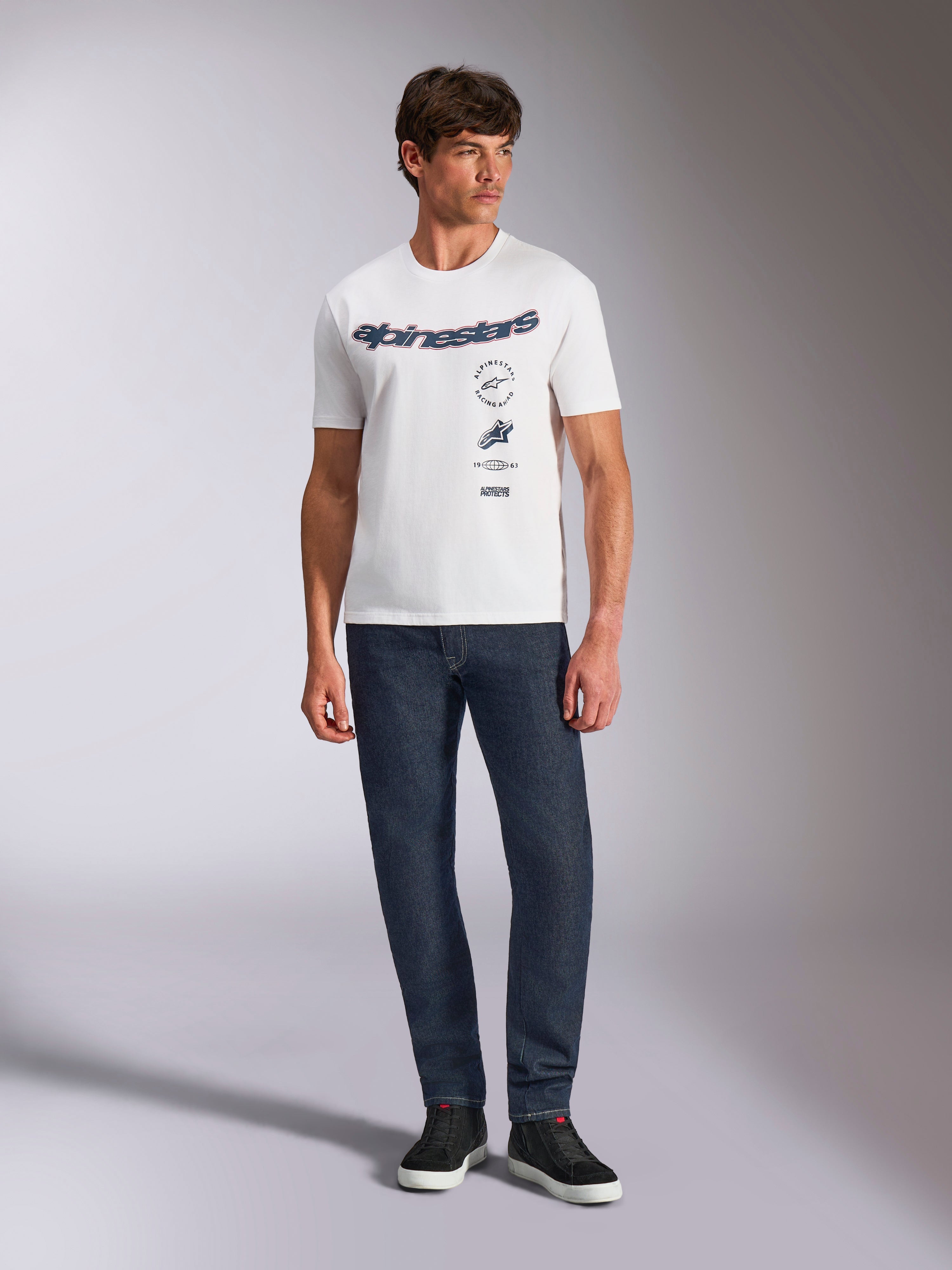 T-shirt Alpinestars Abus CSF, t-shirt à manches courtes, blanc, arborant le logo incurvé Alpinestars et des graphismes superposés sur la poitrine, porté par un mannequin homme avec un jean bleu foncé et des baskets noires, style streetwear décontracté inspiré du sport automobile