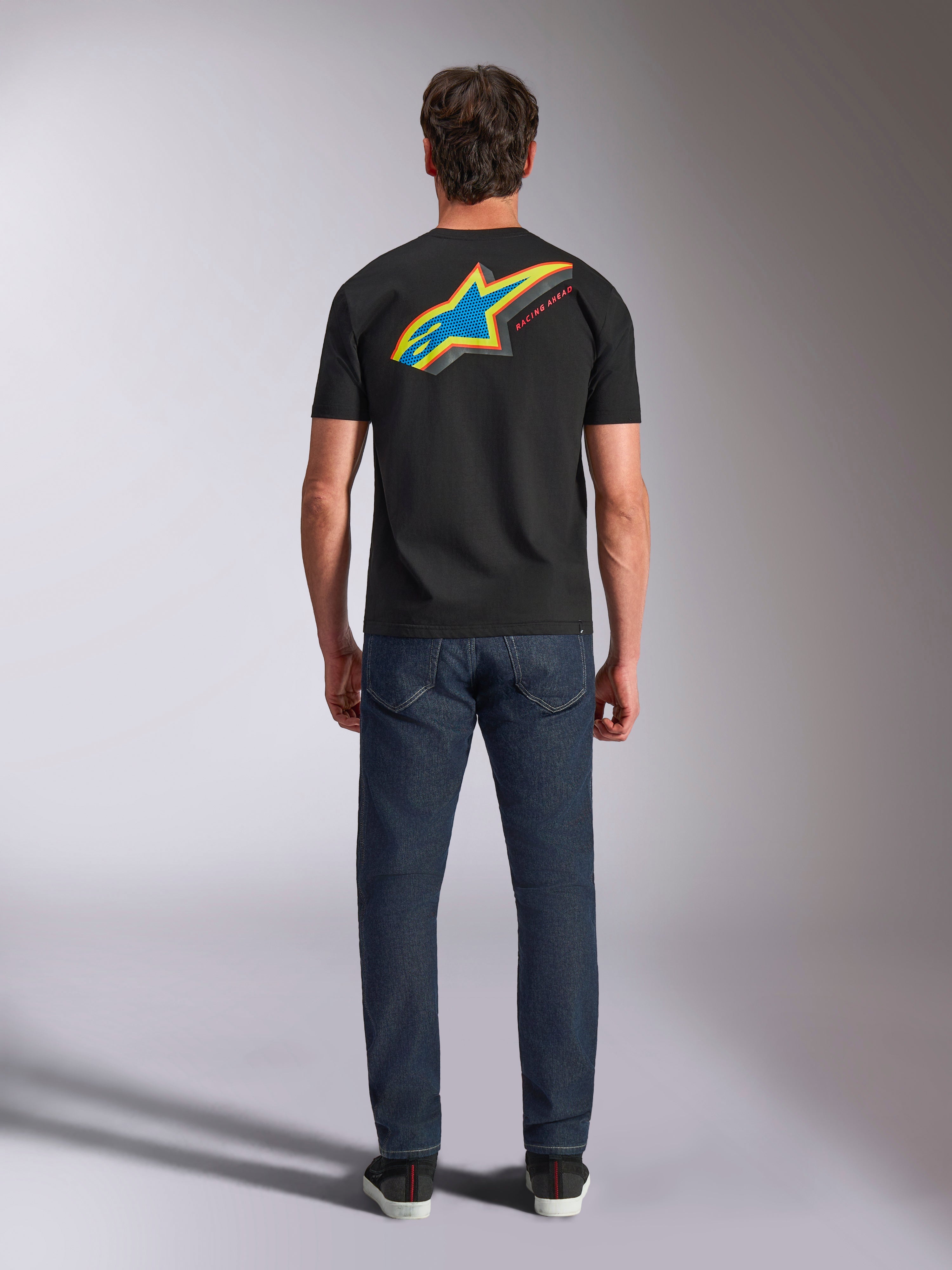 T-shirt Alpinestars Angler CSF, t-shirt à manches courtes, noir, vue de dos du mannequin portant le t-shirt avec un grand logo étoilé Astars multicolore vibrant et le texte « RACING AHEAD », associé à un jean bleu foncé, streetwear décontracté inspiré du sport automobile