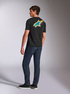 T-shirt Alpinestars Angler CSF, t-shirt à manches courtes, noir, arborant un large logo Astars multicolore éclatant au dos avec des détails perforés bleus, jaunes et rouges, porté avec un jean bleu foncé, style streetwear casual motorsport