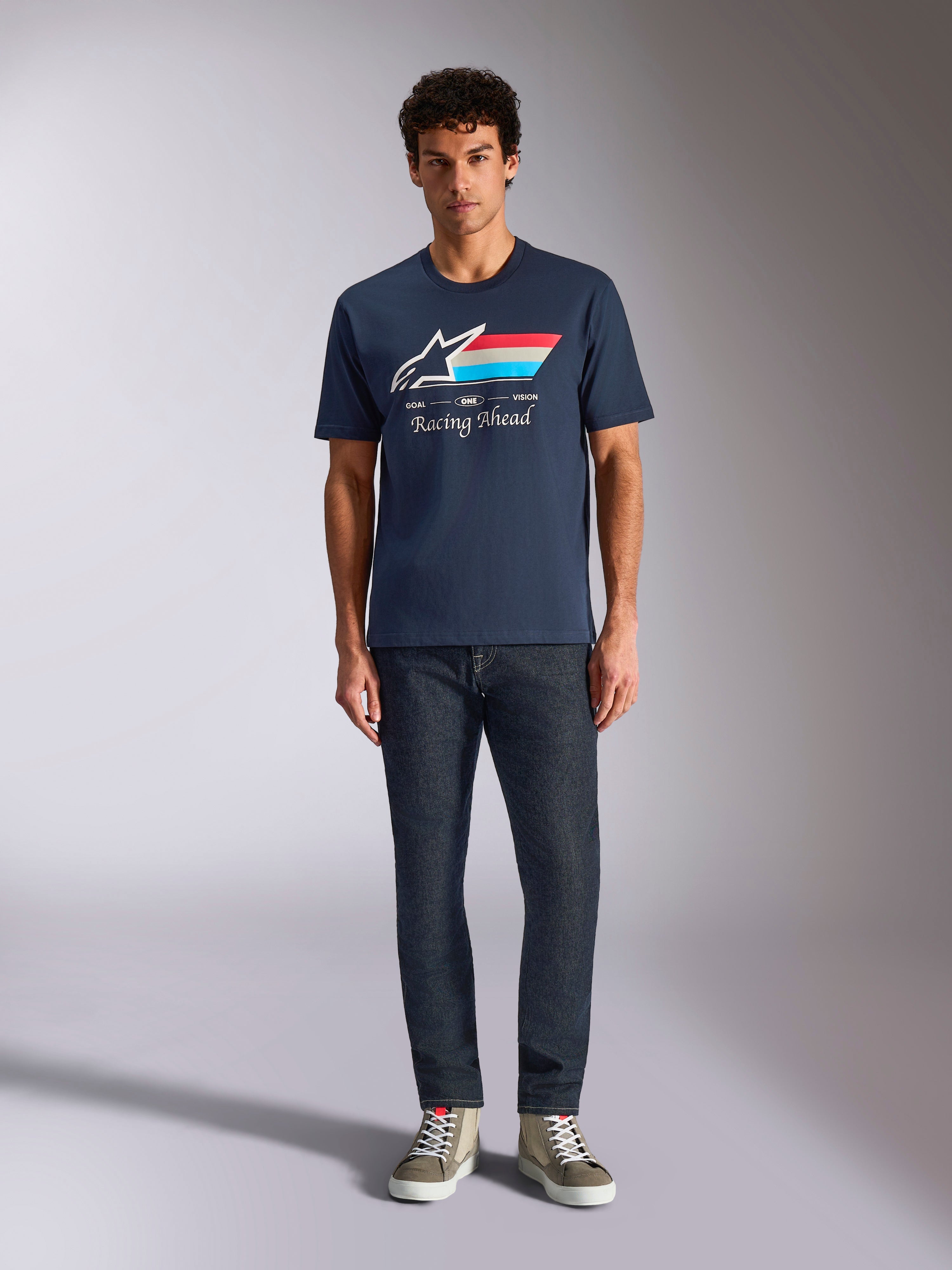 Alpinestars Trifecta CSF Tee, t-shirt à manches courtes, bleu marine, logo Astars blanc avec rayures rouges, blanches et bleu clair et texte « Racing Ahead », porté avec un jean foncé et des baskets olive, style streetwear motorsport décontracté