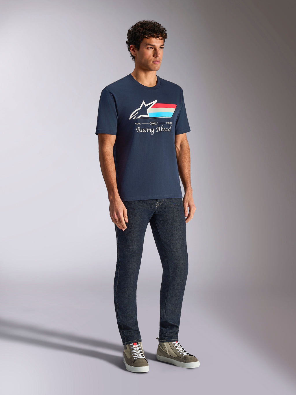 T-shirt Alpinestars Trifecta CSF, manches courtes, bleu marine, avec logo Astars, rayures tricolores et texte « Racing Ahead », porté par un mannequin avec un jean foncé et des baskets, style streetwear décontracté inspiré du sport automobile