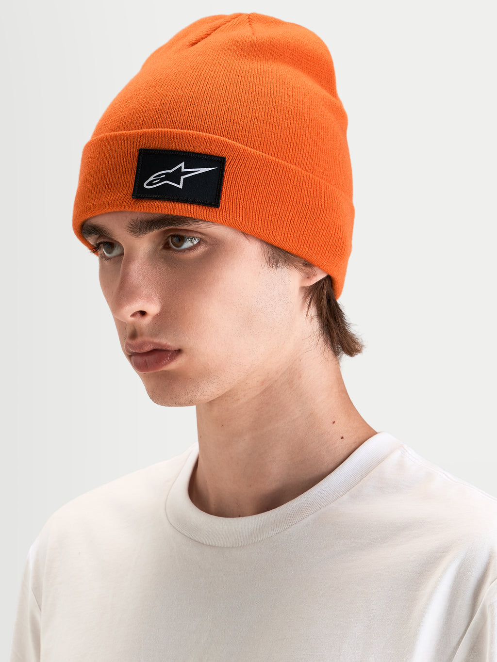 Alpinestars Bonnet Augment Cuff, bonnet sportswear orange en maille, avec un écusson tissé du logo Astars noir et blanc, porté par un modèle dans un style décontracté