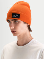 Alpinestars Bonnet Augment Cuff, bonnet sportswear orange en maille, avec un écusson tissé du logo Astars noir et blanc, porté par un modèle dans un style décontracté