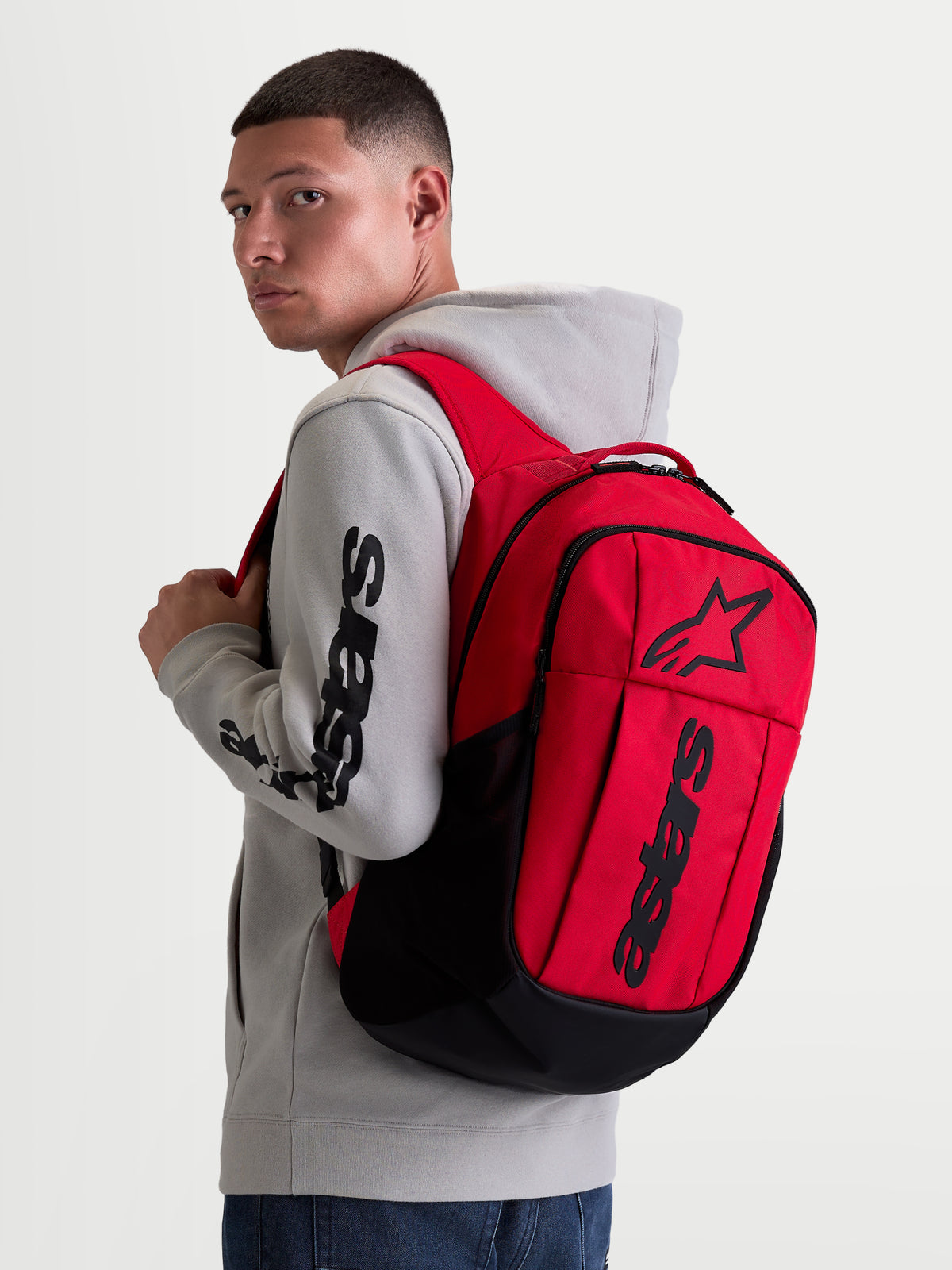 Alpinestars Sac à dos GFX Boss, sac à dos sportswear de 20 litres, rouge et noir, porté par un modèle avec un sweat à capuche gris. Conception en polyester 600D durable avec logo Astars en silicone en relief, idéal pour une utilisation quotidienne urbaine.