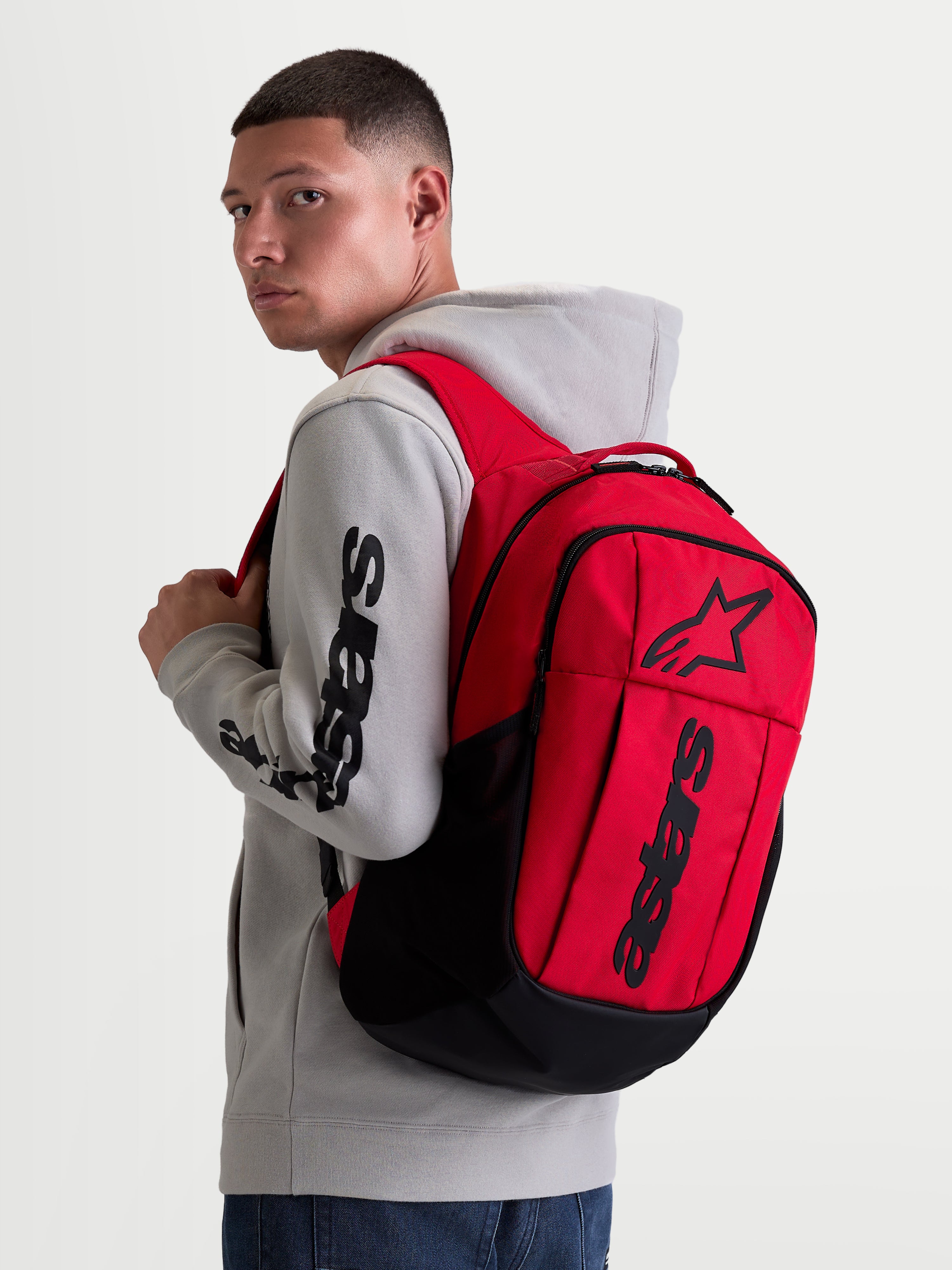 Alpinestars GFX Boss Backpack, 20L Urban Sportswear Daypack, Red and Black, shown on model wearing a grey hoodie. Featuring durable 600D polyester construction, raised silicone Astars graphic branding, and internal laptop sleeve. Ideal for daily commuting and everyday carry.