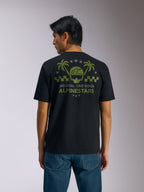 Alpinestars Order CSF Tee, t-shirt à manches courtes, noir, vue de dos, graphisme vert olive avec palmiers et logo de casque, texte 'ONE GOAL. ONE VISION. ALPINESTARS', porté avec un jean bleu, style streetwear décontracté