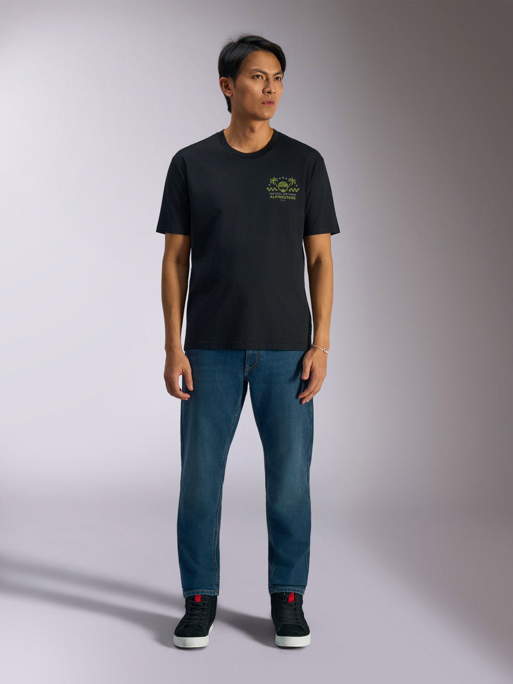 T-shirt Alpinestars Order CSF, t-shirt à manches courtes, noir, motif graphique vert citron avec casque et palmiers sur la poitrine, porté avec un jean bleu et des baskets noires, streetwear motorsport décontracté