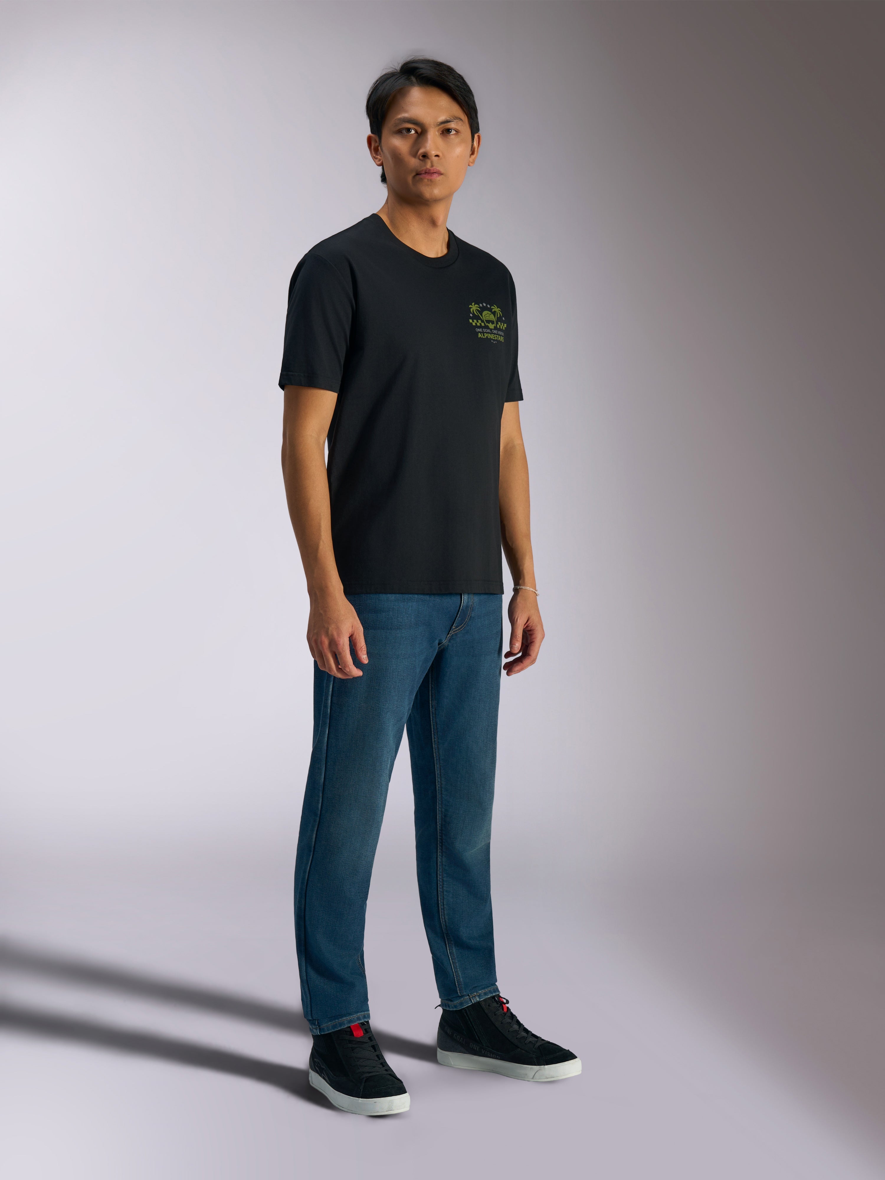 Alpinestars Order CSF Tee, t-shirt à manches courtes, noir, avec logos verts représentant un casque et des palmiers sur la poitrine, style streetwear décontracté, porté avec un jean bleu et des baskets noires