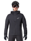 Veste imperméable Alpinestars A-Dura Elite, noire, veste de VTT, design à col montant avec capuche, fermeture éclair frontale avec rabat de protection et logo blanc, portée avec un casque noir et des gants de cyclisme, protection technique pour le trail