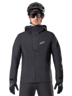 Veste imperméable Alpinestars A-Dura Elite, noire, veste de VTT, design à col montant avec capuche, fermeture éclair frontale avec rabat de protection et logo blanc, portée avec un casque noir et des gants de cyclisme, protection technique pour le trail