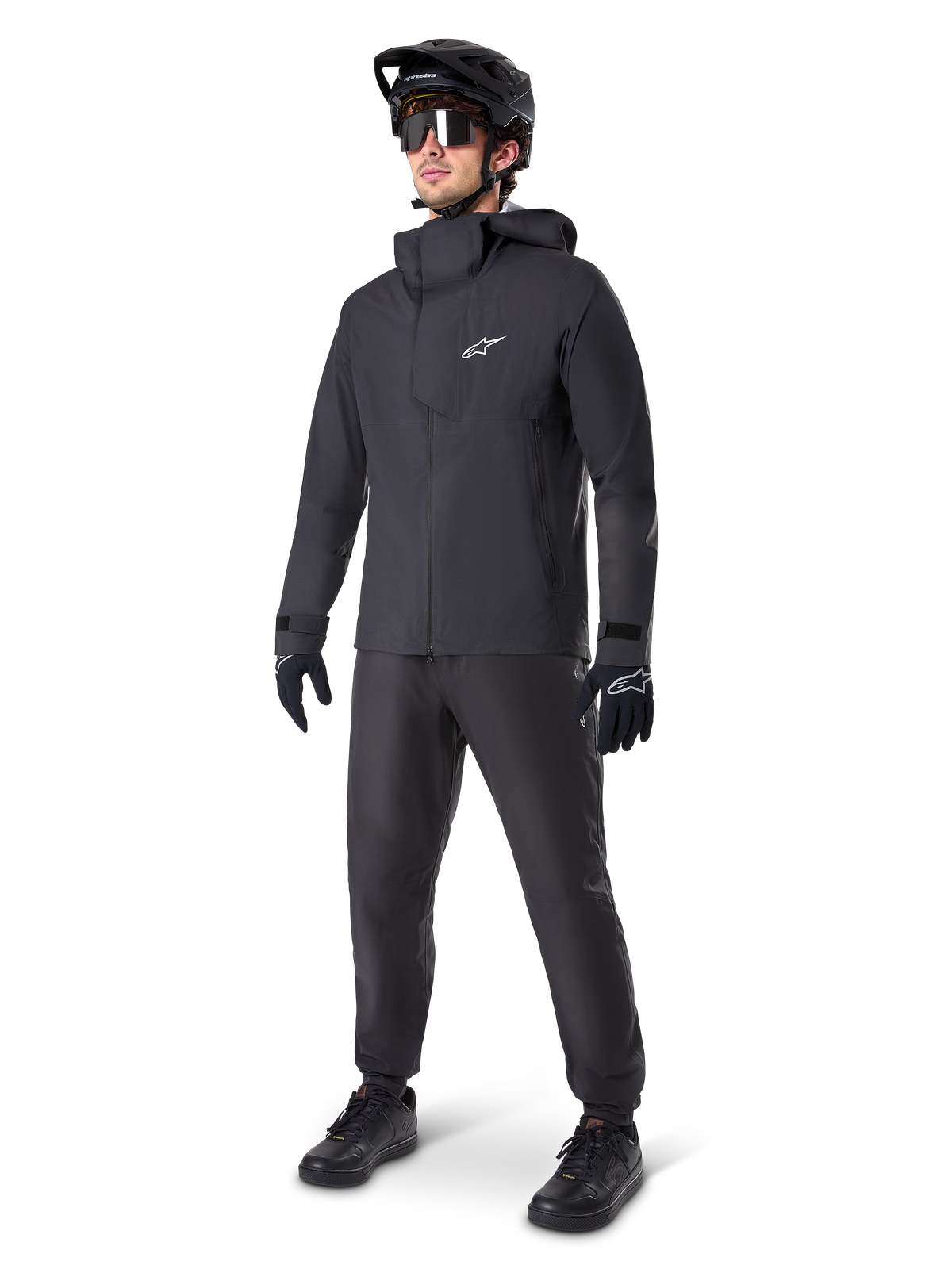 Veste imperméable Alpinestars A-Dura Elite en noir, portée par un pilote avec pantalon de VTT, gants et casque noirs assortis. La veste présente une conception technique imperméable avec un logo Astars blanc sur la poitrine et un col montant protecteur.