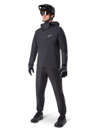 Veste imperméable Alpinestars A-Dura Elite en noir, portée par un pilote avec pantalon de VTT, gants et casque noirs assortis. La veste présente une conception technique imperméable avec un logo Astars blanc sur la poitrine et un col montant protecteur.