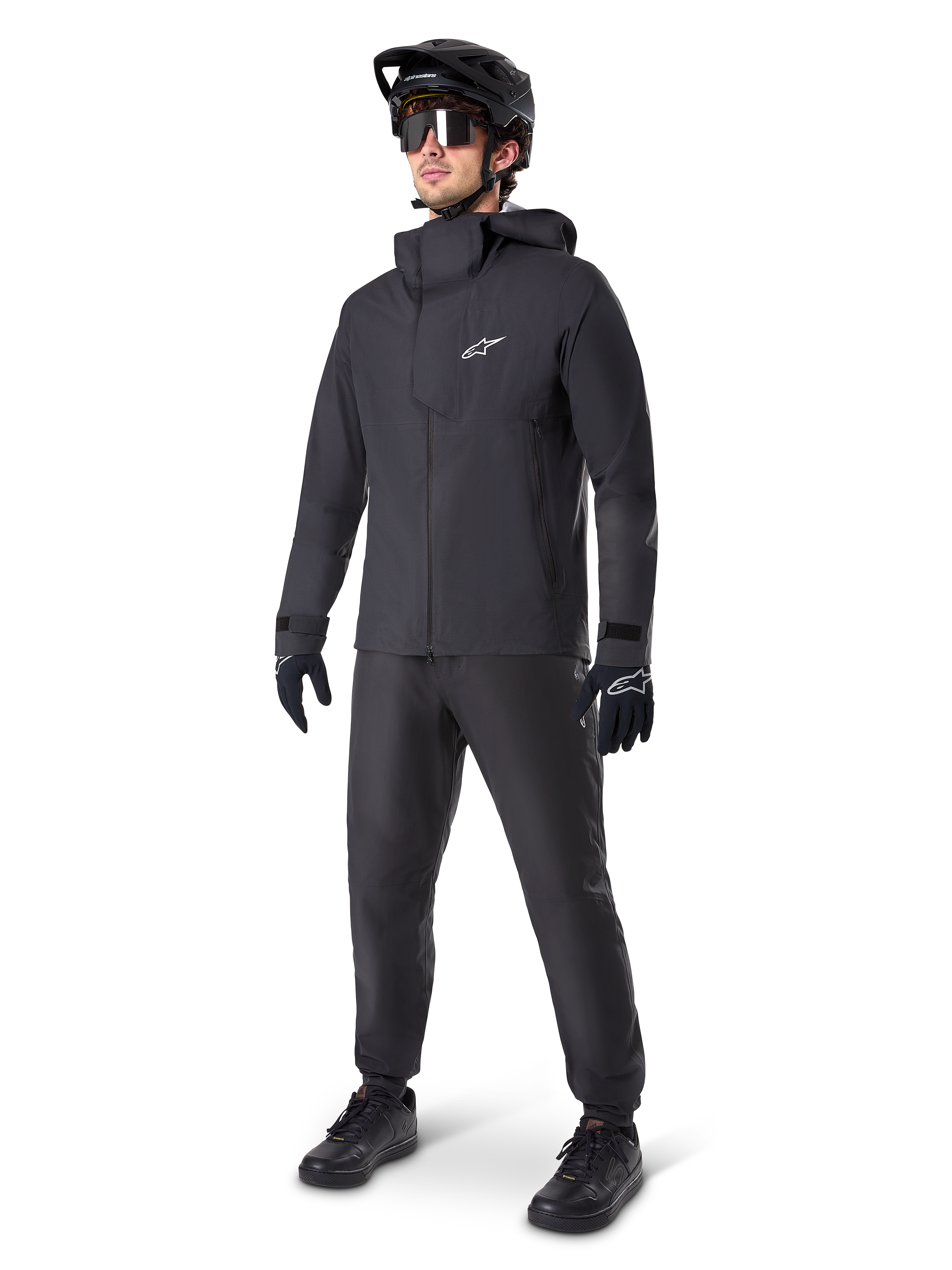 Veste imperméable Alpinestars A-Dura Elite en noir, portée par un pilote avec pantalon de VTT, gants et casque noirs assortis. La veste présente une conception technique imperméable avec un logo Astars blanc sur la poitrine et un col montant protecteur.