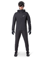 Veste imperméable Alpinestars A-Dura Elite, noire, portée avec un pantalon et des gants de cyclisme assortis, casque noir et lunettes de soleil, logo Astars blanc sur la poitrine, design technique haute performance pour le VTT