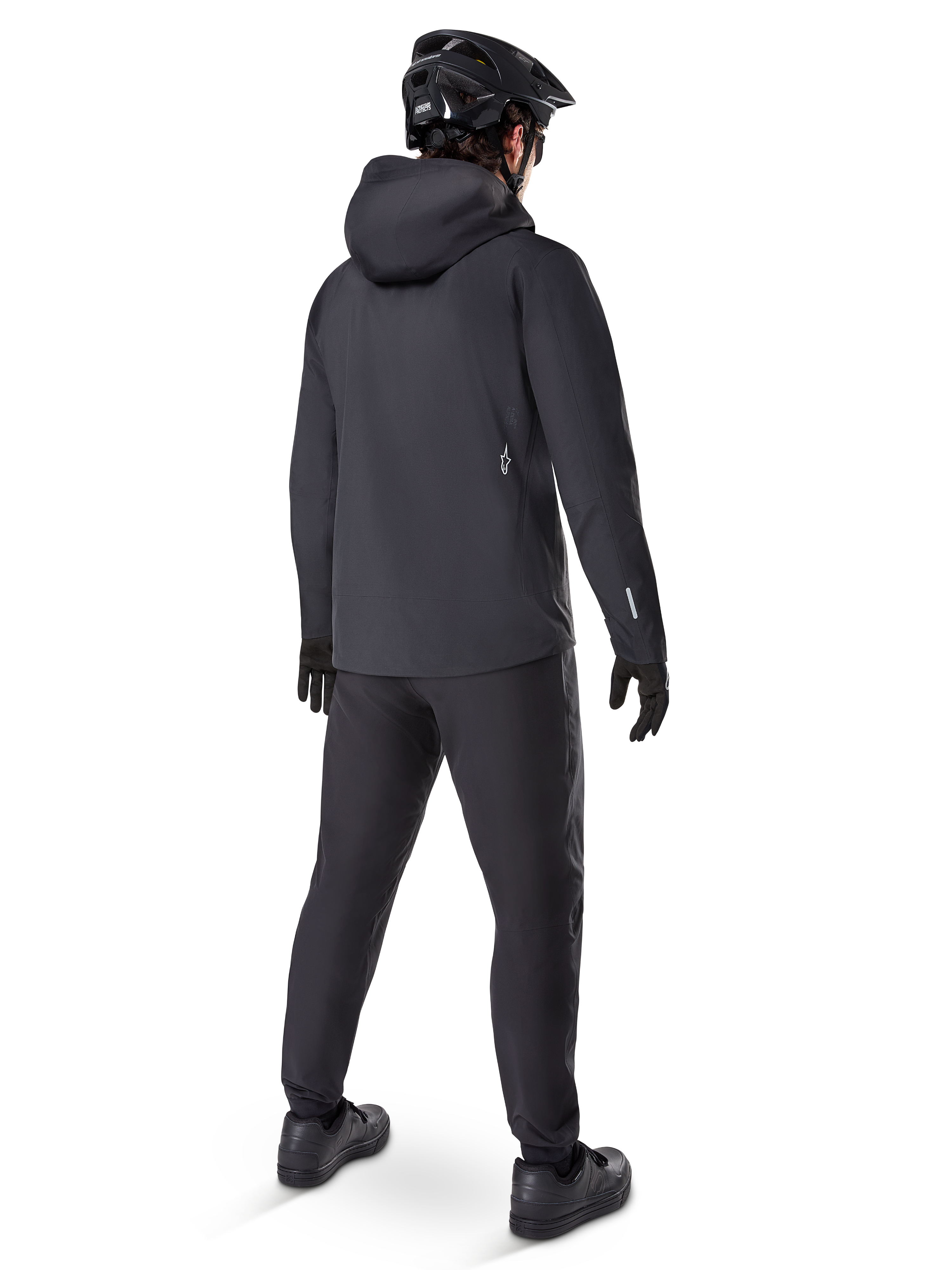 Veste imperméable Alpinestars A-Dura Elite, noire, vue de dos montrant la capuche par-dessus le casque et l'ourlet arrière plus long, associée à un pantalon de VTT, des gants et un casque noirs assortis pour une protection complète sur les sentiers par tous les temps.