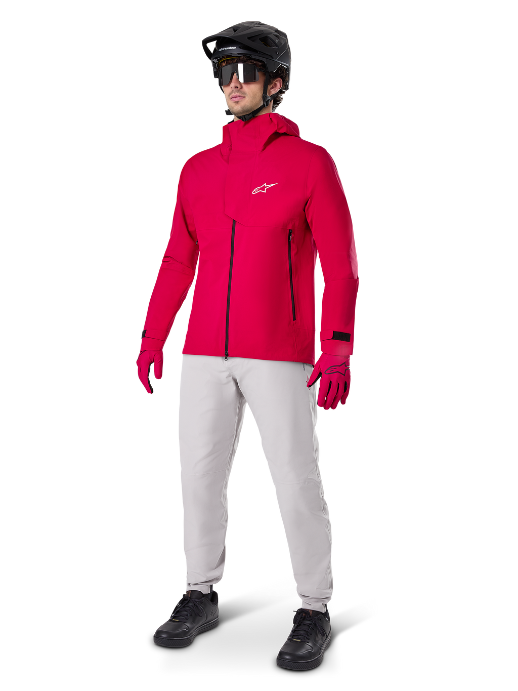 Cycliste portant la veste imperméable Alpinestars A-Dura Elite en Rouge Fluo, associée à un pantalon technique gris clair, des gants rouges, un casque de VTT noir avec lunettes et des chaussures noires, tenue haute performance complète pour le VTT