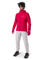 Cycliste portant la veste imperméable Alpinestars A-Dura Elite en Rouge Fluo, associée à un pantalon technique gris clair, des gants rouges, un casque de VTT noir avec lunettes et des chaussures noires, tenue haute performance complète pour le VTT