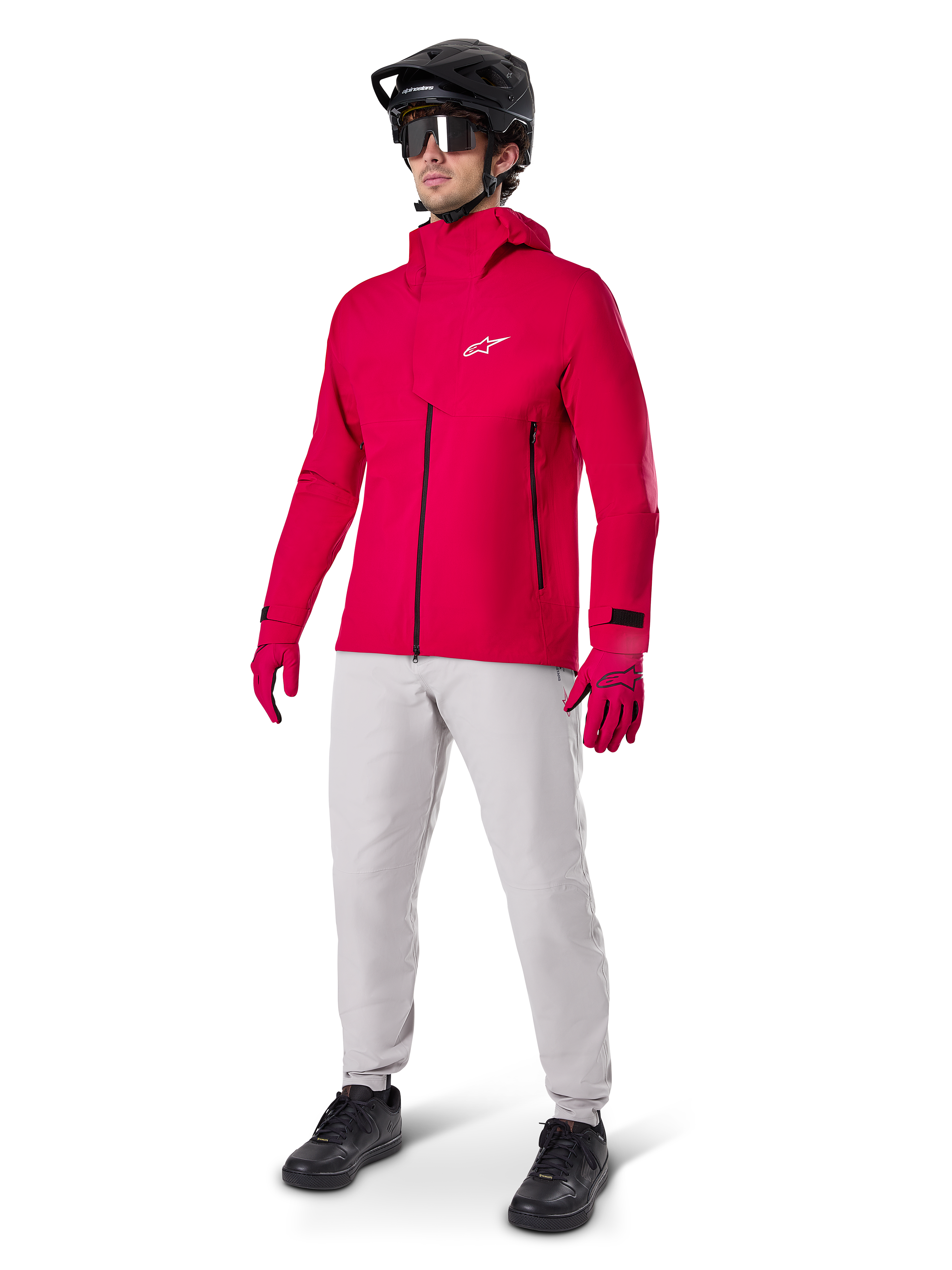 Cycliste portant la veste imperméable Alpinestars A-Dura Elite en Rouge Fluo, associée à un pantalon technique gris clair, des gants rouges, un casque de VTT noir avec lunettes et des chaussures noires, tenue haute performance complète pour le VTT