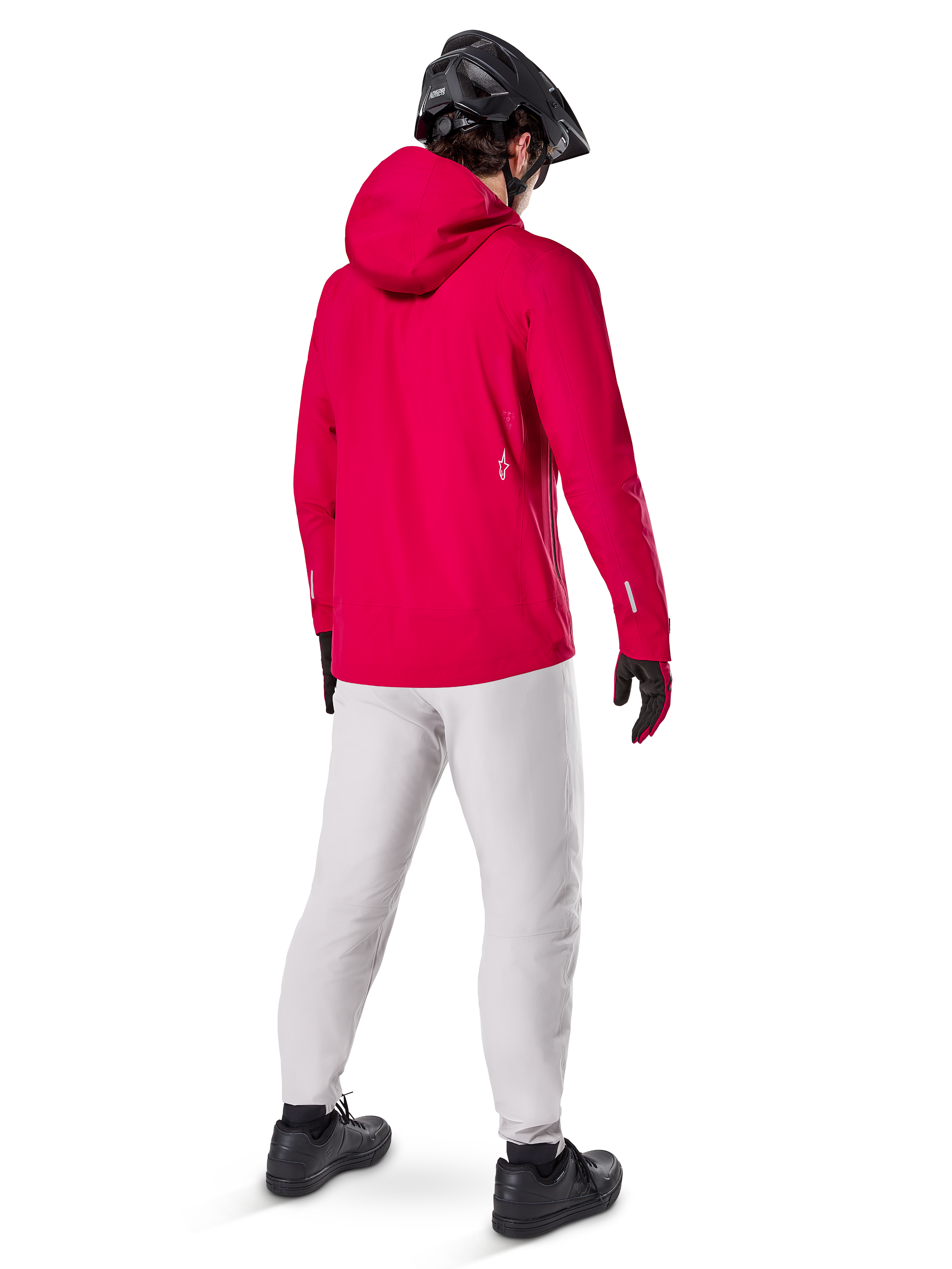 Veste imperméable Alpinestars A-Dura Elite, rouge fluo, vue de dos de trois-quarts, portée avec un pantalon gris clair et un casque de VTT noir, dotée d'une capuche et de détails réfléchissants, veste de pluie technique haute performance pour le trail