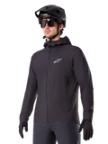 Alpinestars A-Dura Thermal Jacket, Black Cycling Jacket, hooded design with front zipper, white star logo on chest, worn with black mountain bike helmet and gloves, sporty performance gear for cold weather trail riding