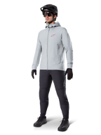 Veste thermique Alpinestars A-Dura en Titanium, veste de cyclisme à capuche gris clair avec logo Alpinestars rouge sur la poitrine, portée par un homme avec casque de VTT noir, lunettes de soleil, pantalon gris foncé et gants noirs, équipement de VTT professionnel