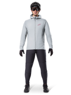 Veste thermique Alpinestars A-Dura, gris titane, veste de cyclisme portée avec un pantalon de VTT noir, des gants et un casque, dotée d'un petit logo étoile rouge sur la poitrine et d'une fermeture éclair frontale intégrale, équipement isolant léger pour le tout-terrain.